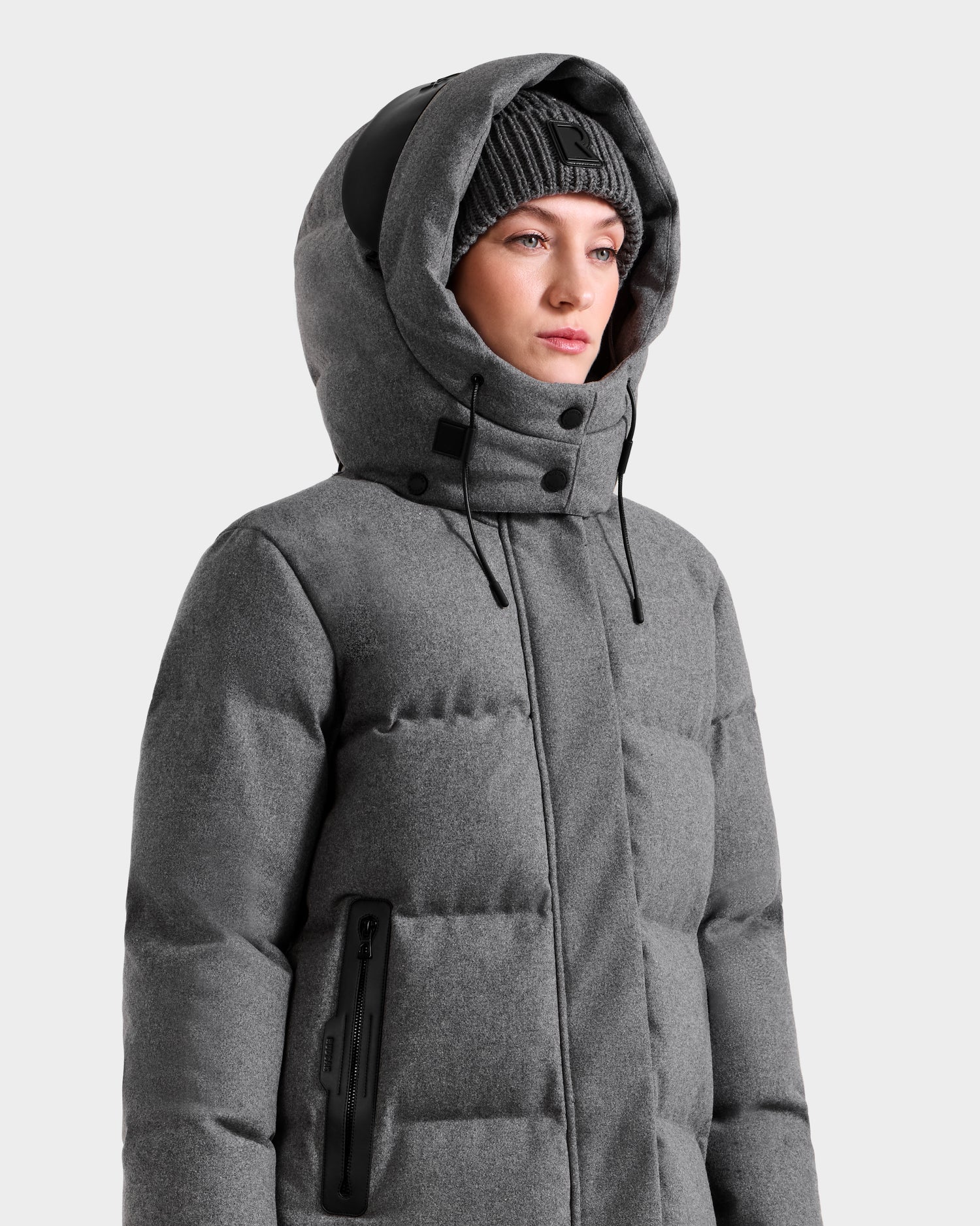 A person wears the Rudsak MAO W - 8125568 Heather Charcoal puffer coat with a hood and a gray beanie, standing against a plain light background and showcasing stylish winter outerwear.