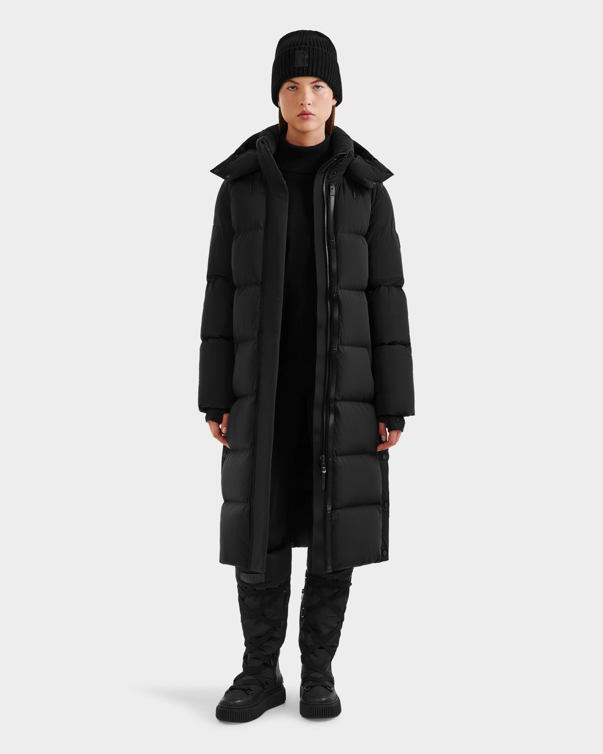 A person stands facing forward wearing the Rudsak MAO - 8124780 Black womens long down coat, paired with a black beanie, pants, and boots—ideal winter wear—against a plain white background.