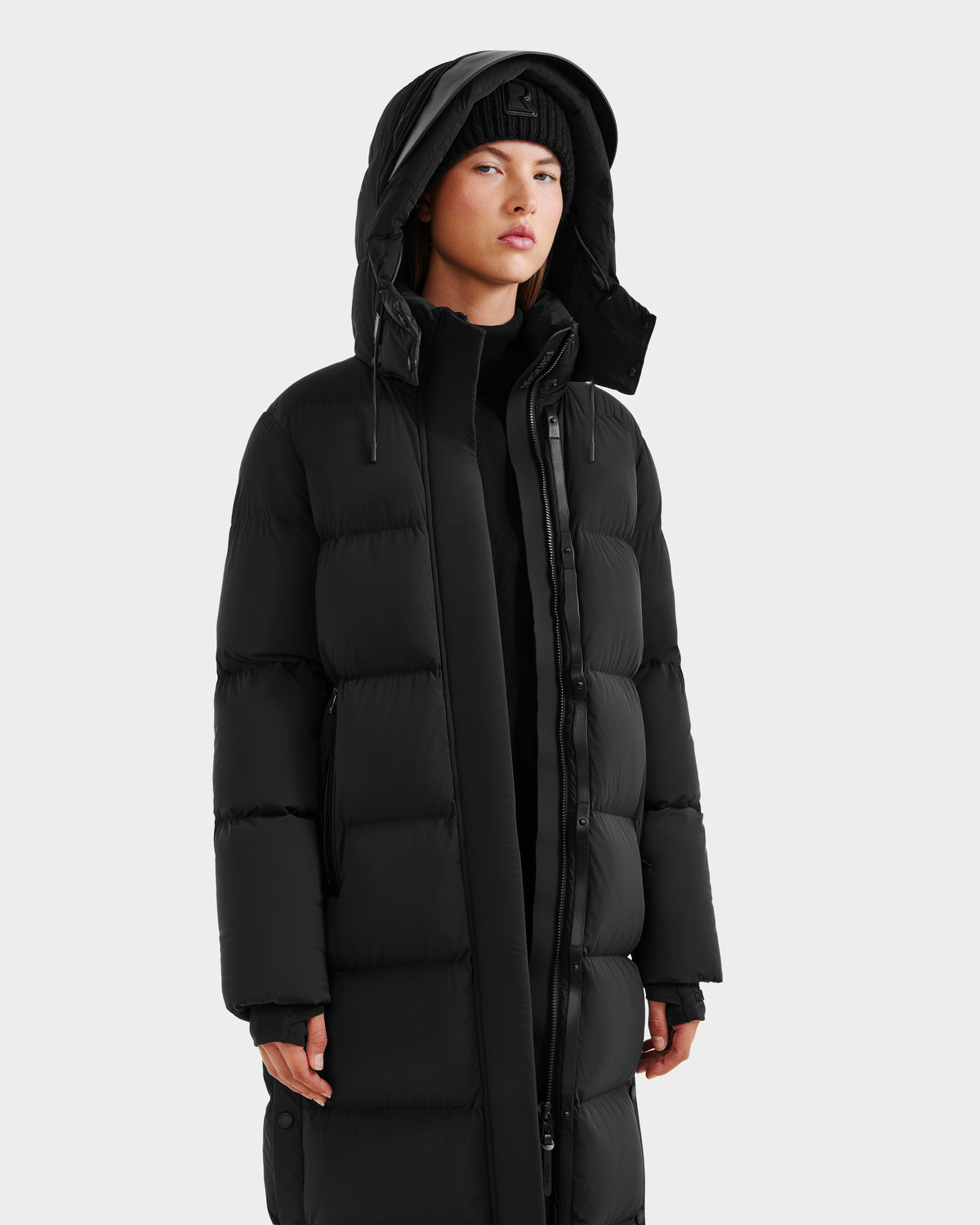 A person wearing the Rudsak MAO - 8124780 Black women’s long down coat with a hood and a black beanie stands against a light gray background—ideal winter wear for chilly days.