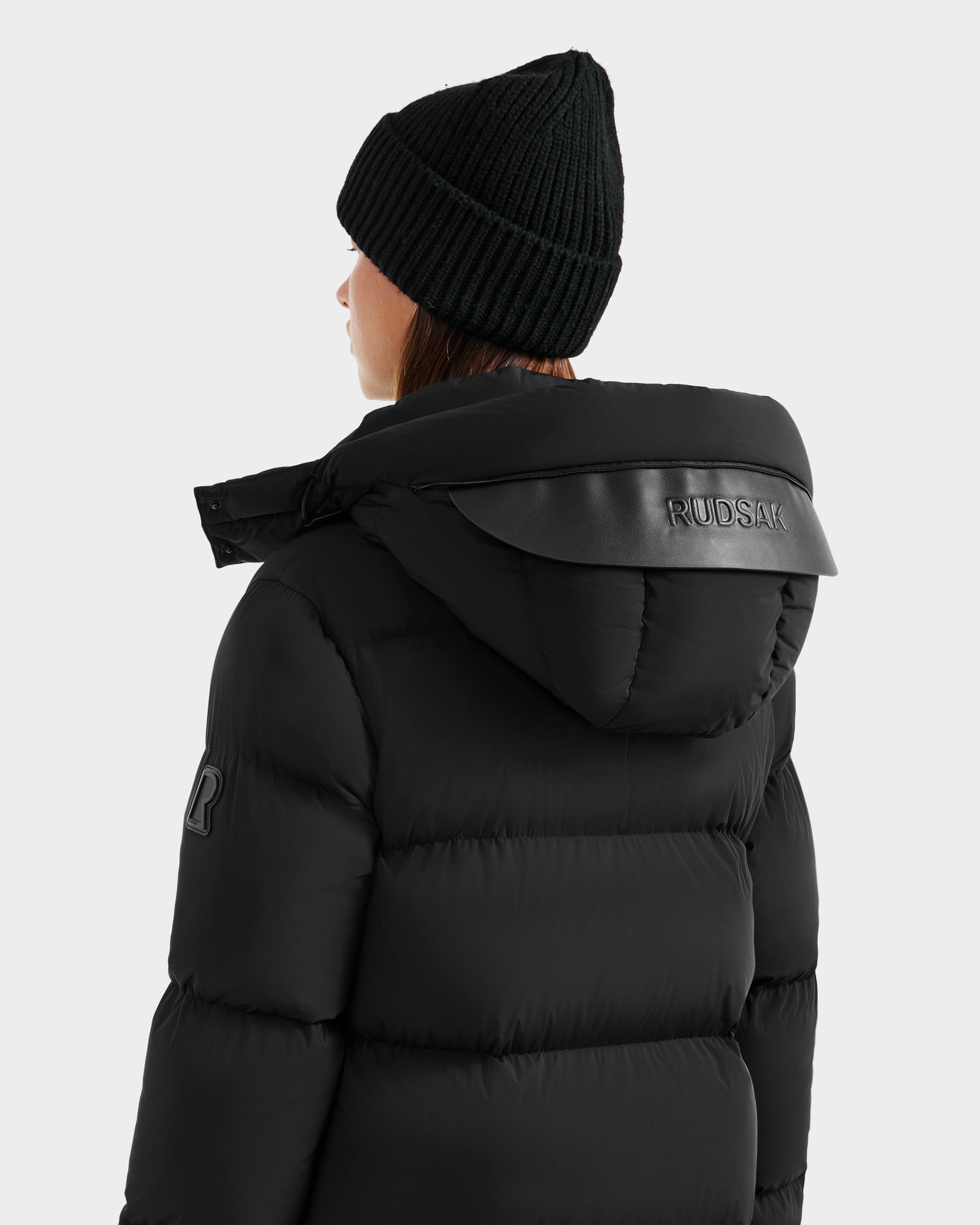 A person wearing the Rudsak MAO - 8124780 Black water-resistant puffer jacket with a hood and a black knit beanie is viewed from behind. The Rudsak logo is visible on the hood and left sleeve, with a plain light gray background.