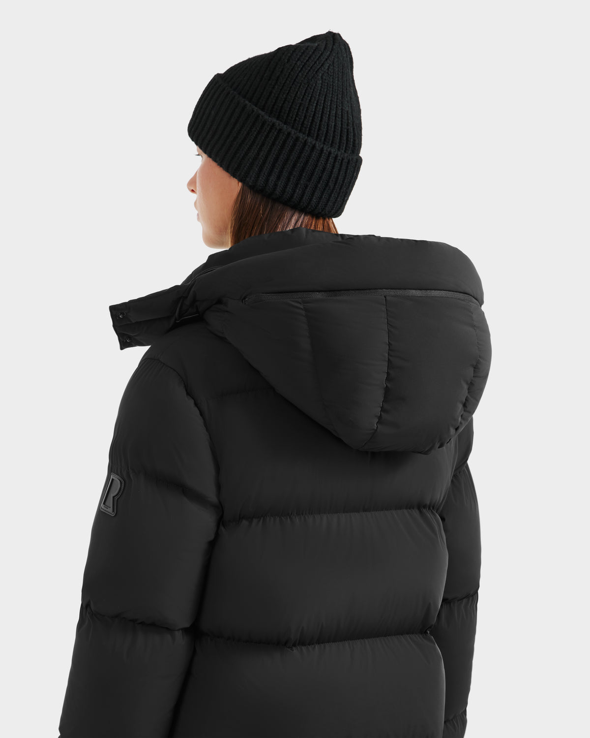 A person in the Rudsak MAO - 8124780 Black water-resistant puffer jacket with a hood and a black knit beanie, seen from behind against a plain light background.