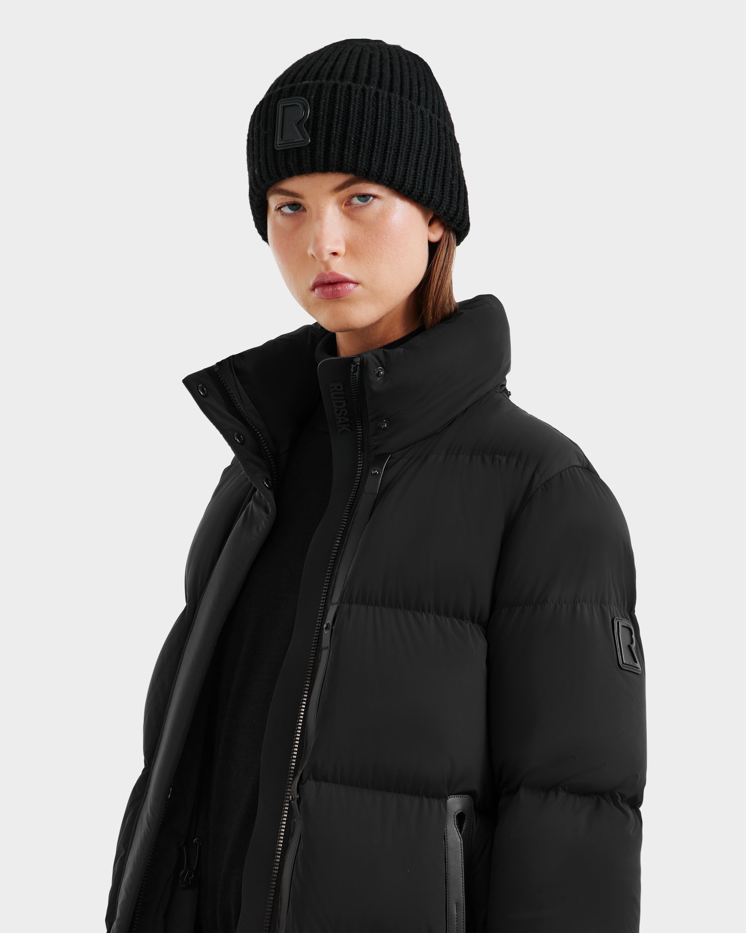 A person wears the Rudsak MAO - 8124780 Black water-resistant puffer jacket and a black knit beanie, standing against a plain light gray background and looking directly at the camera with a neutral expression.