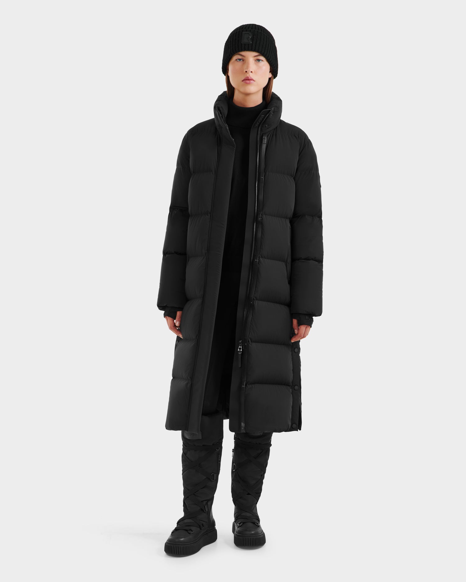 A person stands against a plain background wearing the Rudsak MAO - 8124780 Black womens long down coat, paired with a black beanie, pants, quilted boots, and a turtleneck. Their winter wear is water-resistant; hands by their sides and expression neutral.