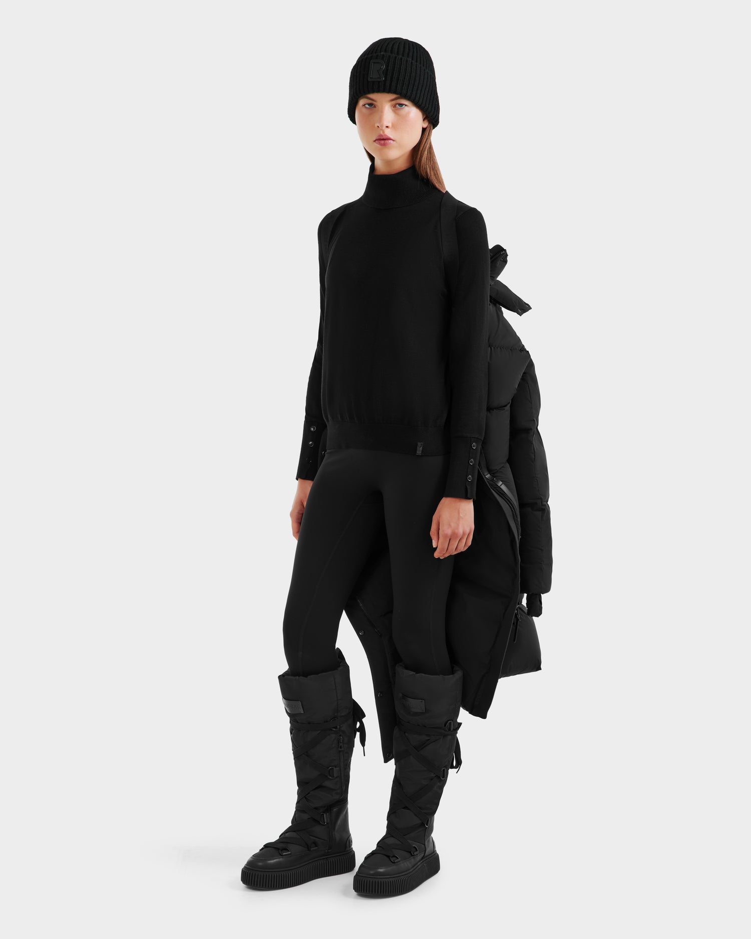 A woman faces forward in a sleek all-black outfit with a beanie, turtleneck, leggings, and the Rudsak MAO - 8124780 Black womens long down coat draped over her shoulders. The background is plain and light-colored.
