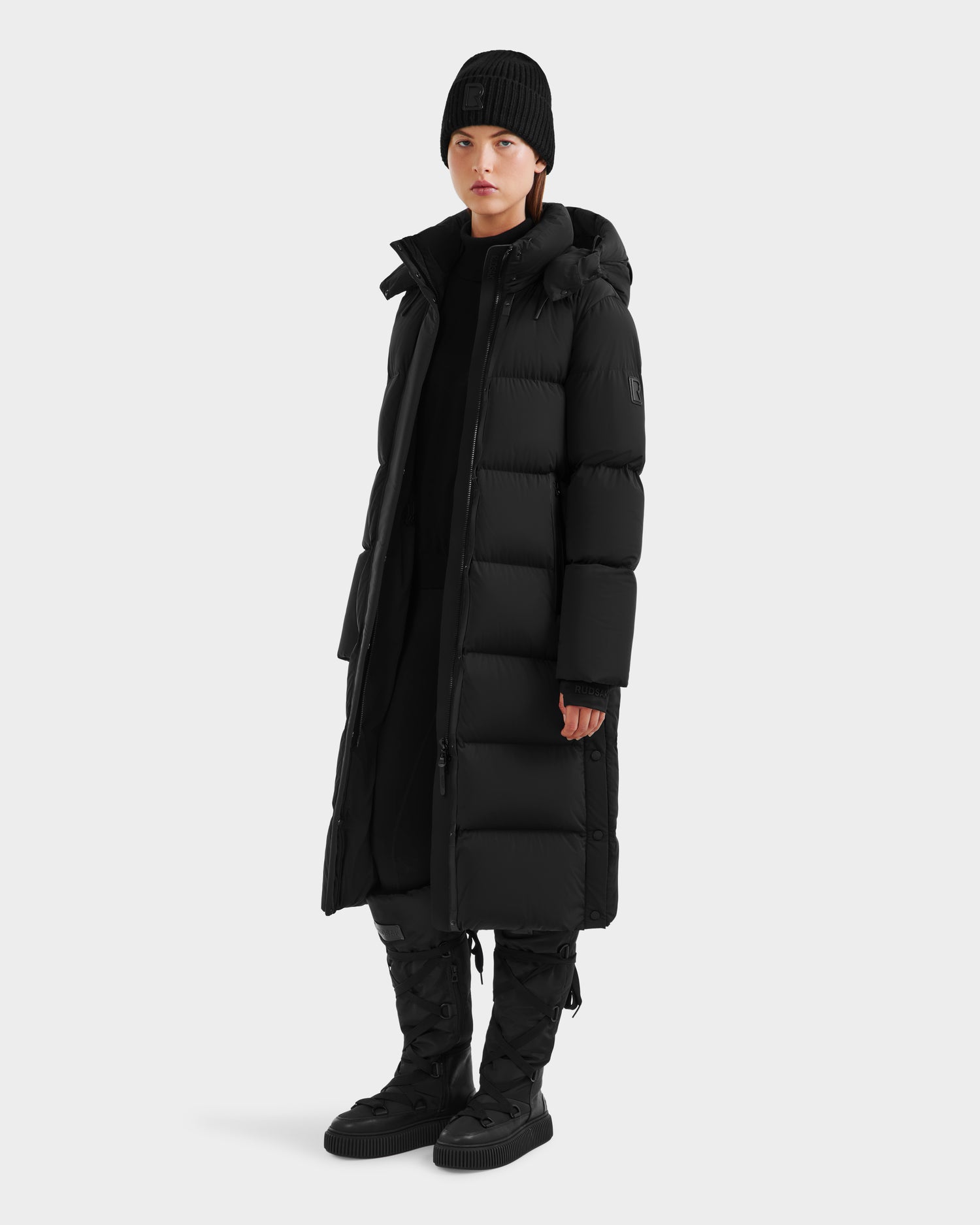 A person faces forward against a plain light gray background, wearing the Rudsak MAO - 8124780 Black womens long down coat—water-resistant winter wear—paired with black pants, winter boots, a beanie, and a top.