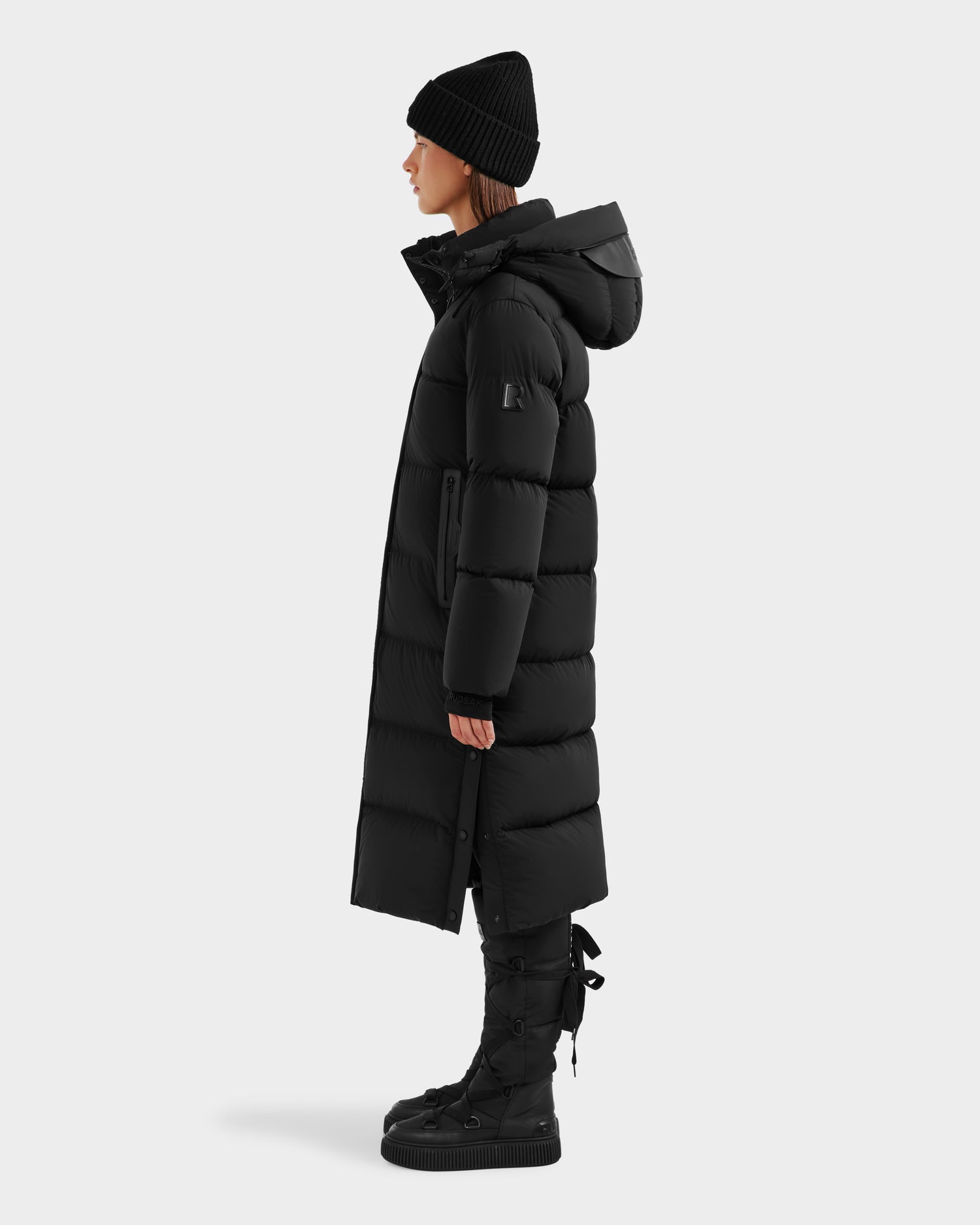 A person in profile models the Rudsak MAO - 8124780 Black womens long, black, water-resistant puffer coat with hood, paired with a black knit beanie and lace-up boots, all set against a plain white background.