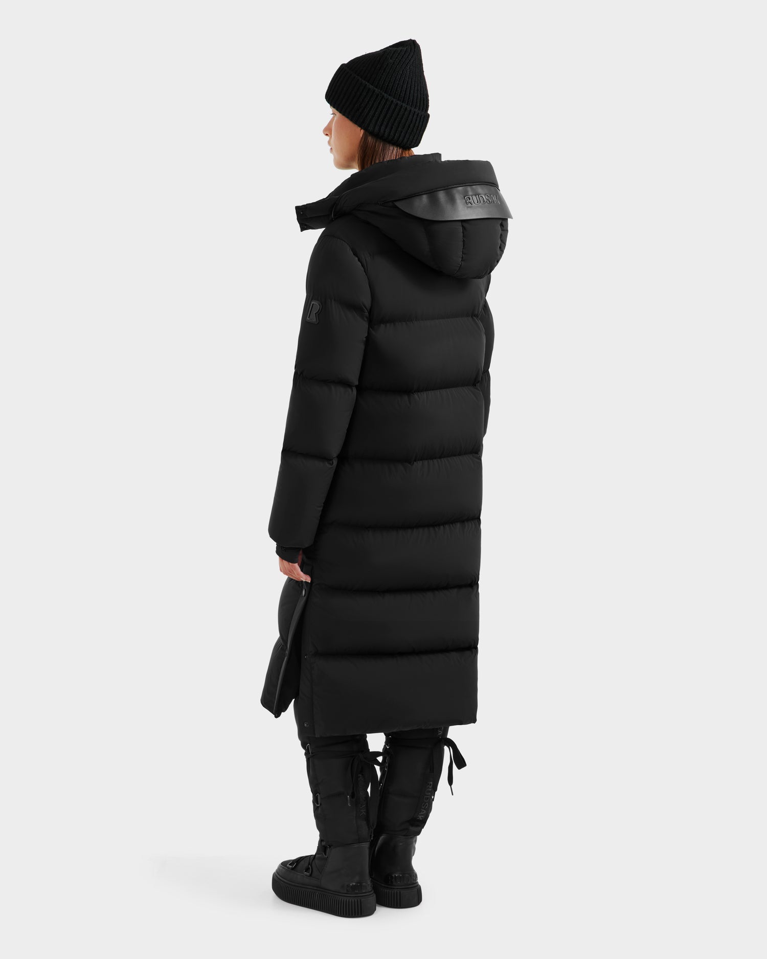 A person in the Rudsak MAO - 8124780 Black women’s water-resistant long down coat, paired with a black beanie, gloves, and boots, stands against a plain light background—a chic showcase of winter style.