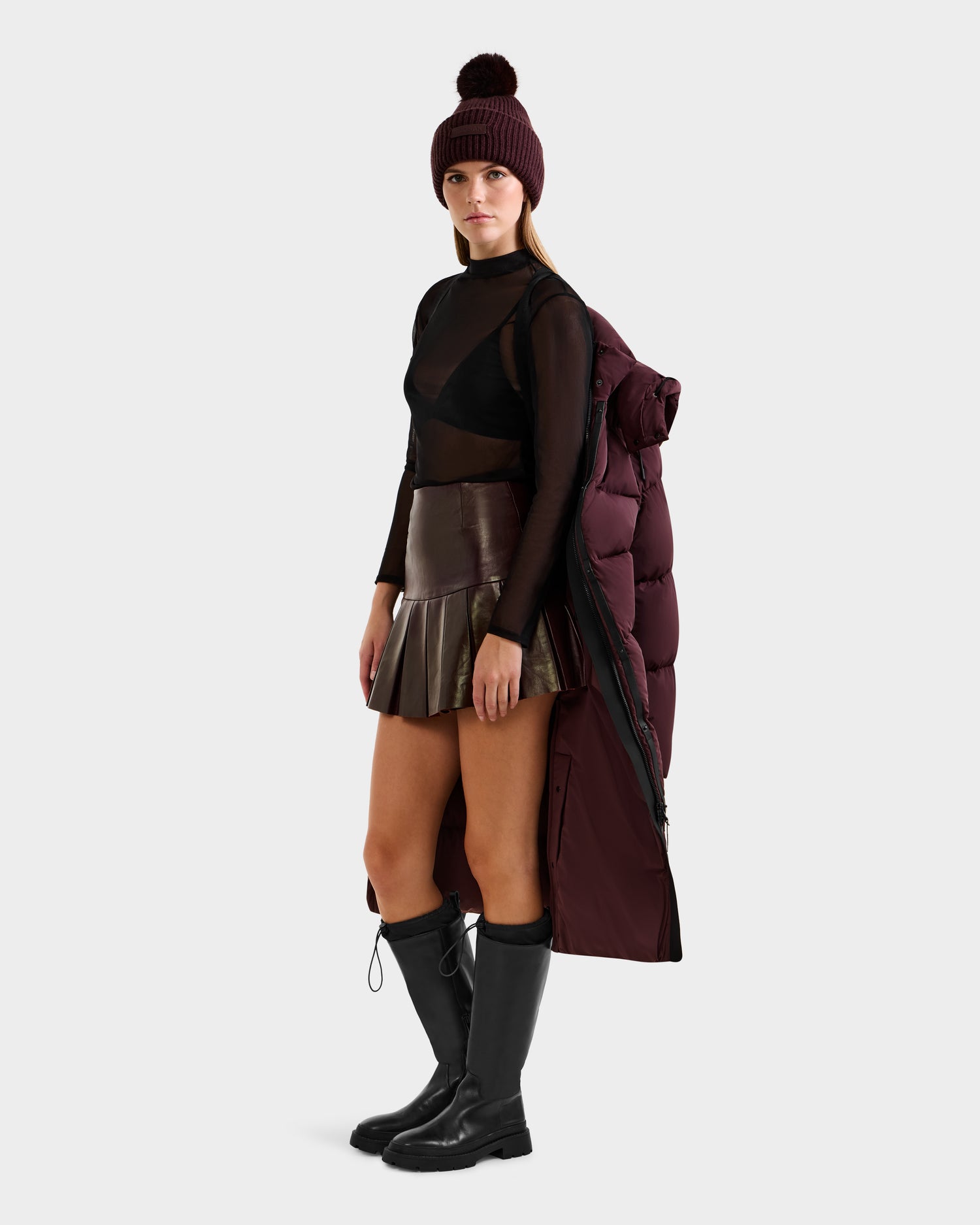 A woman stands confidently against a plain background, draping the Rudsak MAO - 8124780 Bordeaux wind and water resistant maroon puffer jacket over her shoulders, paired with a sheer black top, metallic skirt, boots, and a maroon pom-pom beanie.