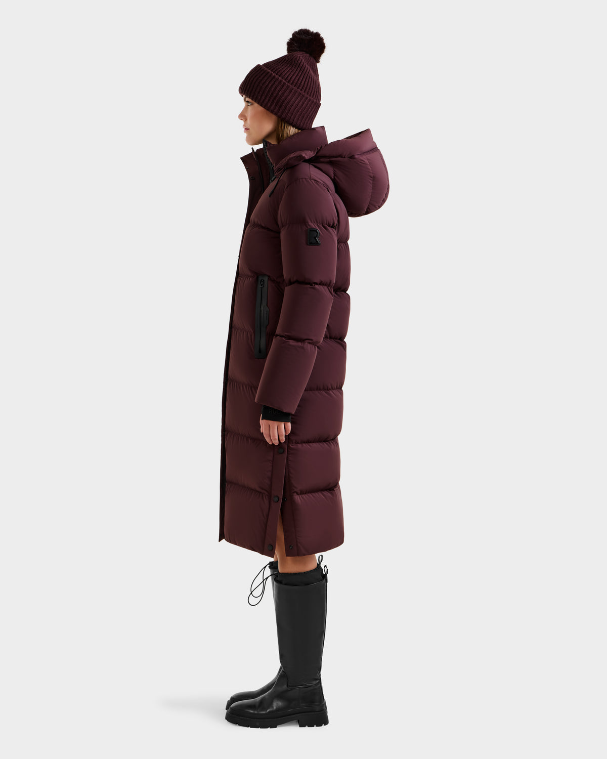 A woman in profile wears the Rudsak MAO - 8124780 Bordeaux women’s long quilted down coat with hood, a maroon knit beanie, and black knee-high boots. The RDS-certified wind- and water-resistant coat is set against a plain white background.