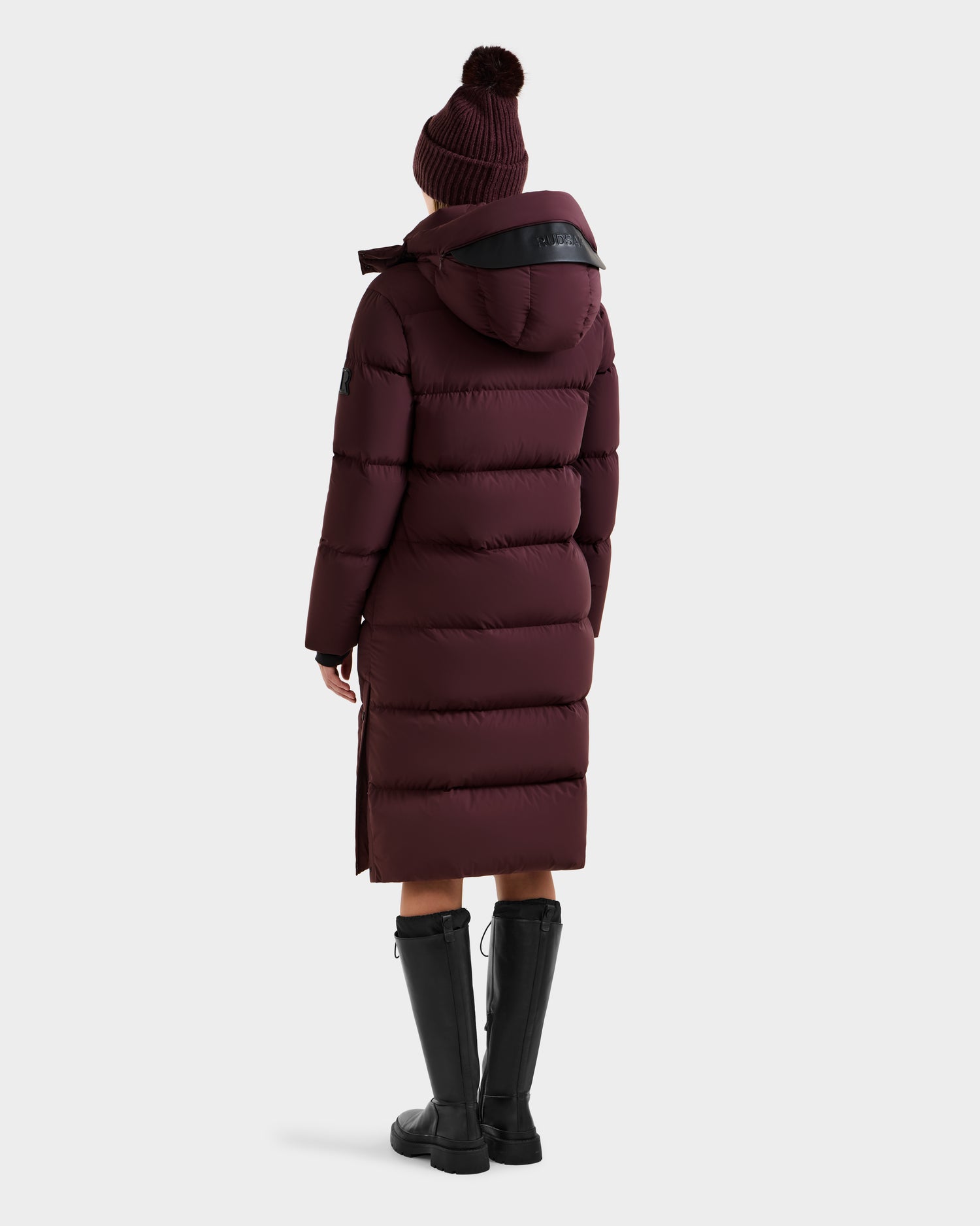 A person wears the Rudsak MAO - 8124780 Bordeaux women's long quilted down coat, wind and water resistant with RDS-certified down, paired with black knee-high boots and a matching maroon hat, standing back to the camera against a plain backdrop.