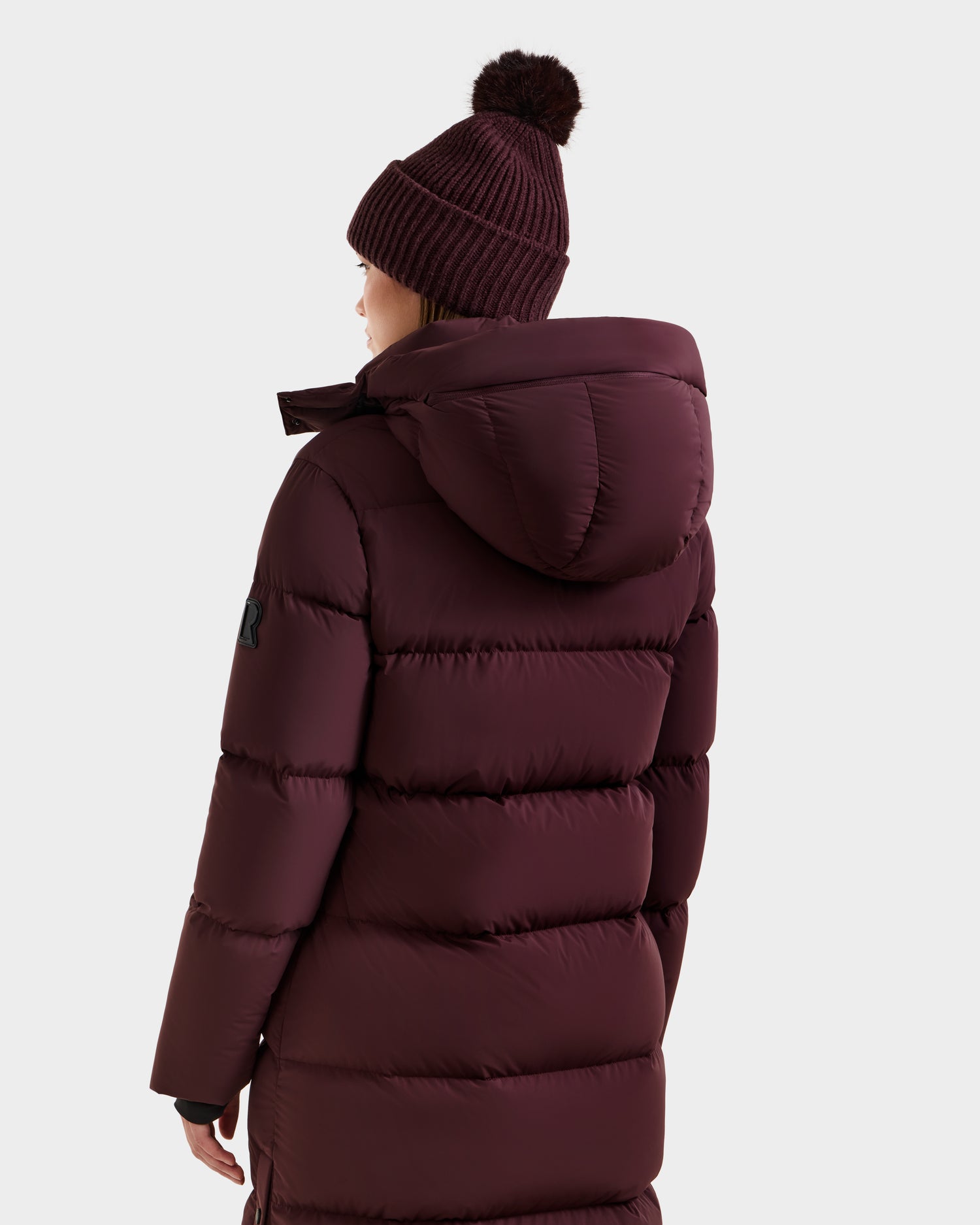 A person wearing the Rudsak MAO - 8124780 Bordeaux women's long quilted down coat with a hood and a matching pom-pom beanie, seen from behind against a plain light background.
