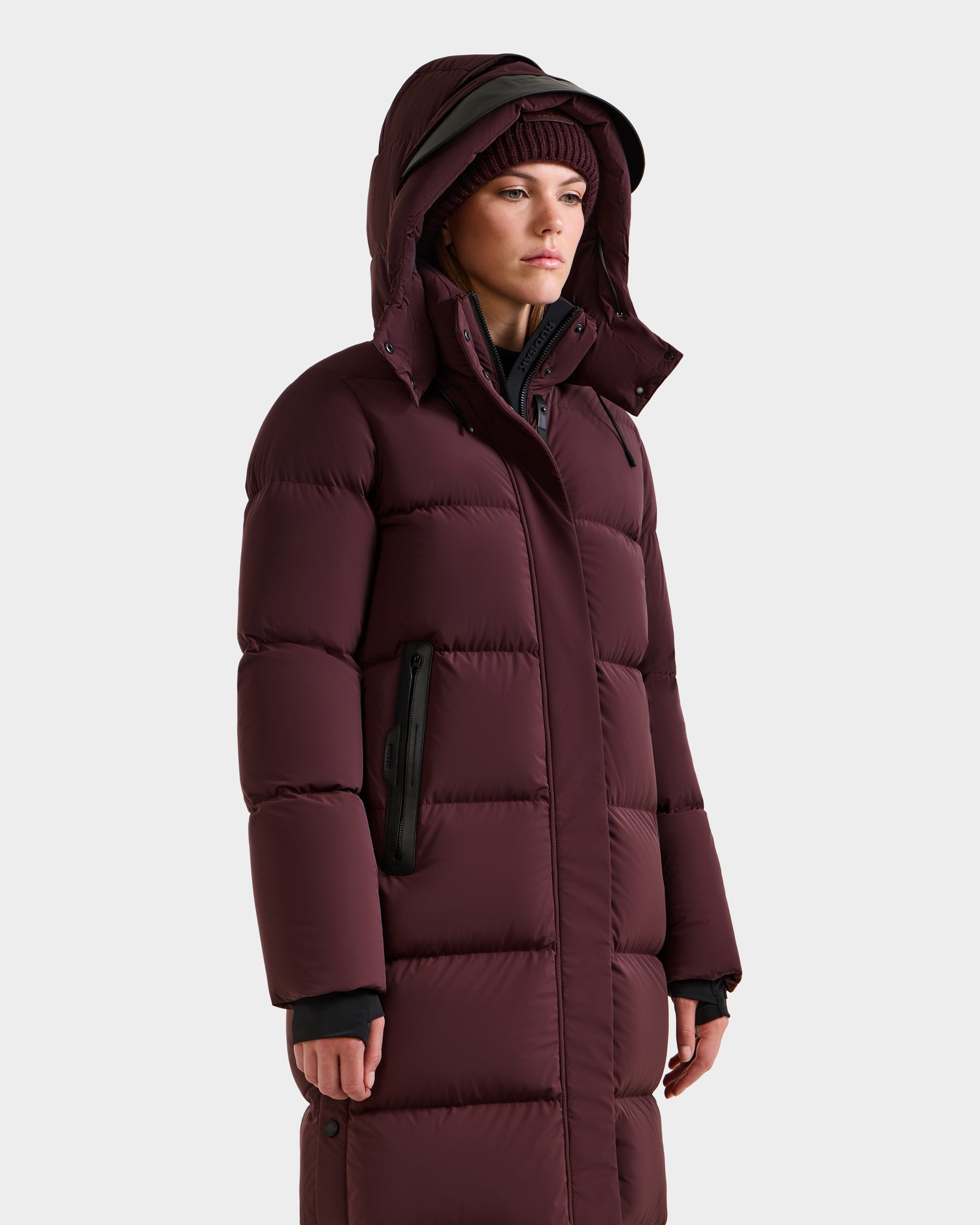 A person wears the Rudsak MAO - 8124780 Bordeaux women’s long quilted down coat with RDS-certified down and black zipper details, paired with a matching knit hat, standing slightly to the side against a plain light gray background.