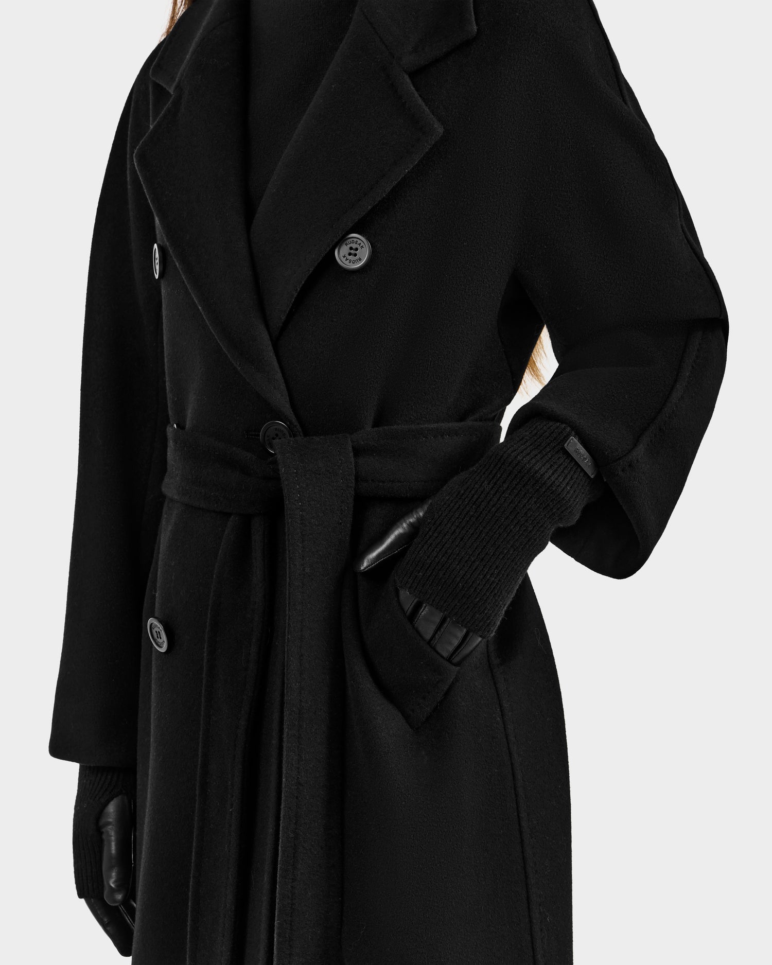 A person wears the Rudsak MARINE - 8424516 Black double-breasted coat with a belt, paired with leather gloves and a black sweater. The cropped image shows only the upper body and arms against a plain background.