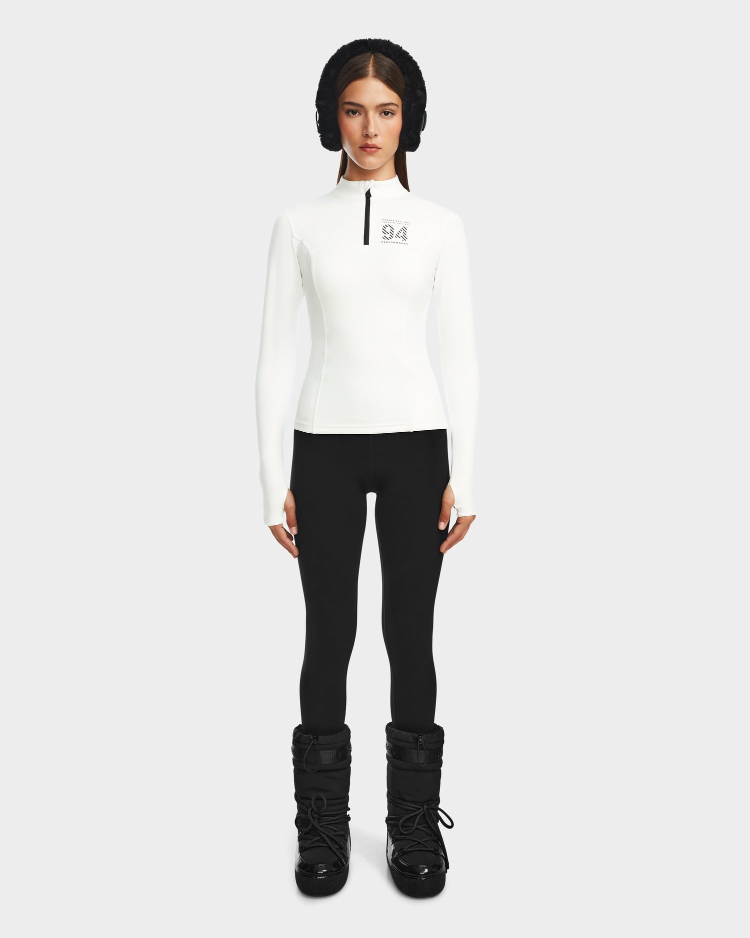 A woman stands facing forward wearing the Rudsak MARIS - 8524603 Optic White womens active zip-up top, styled with black leggings, winter boots, and a headband—an ideal ski layer—against a plain light gray background.