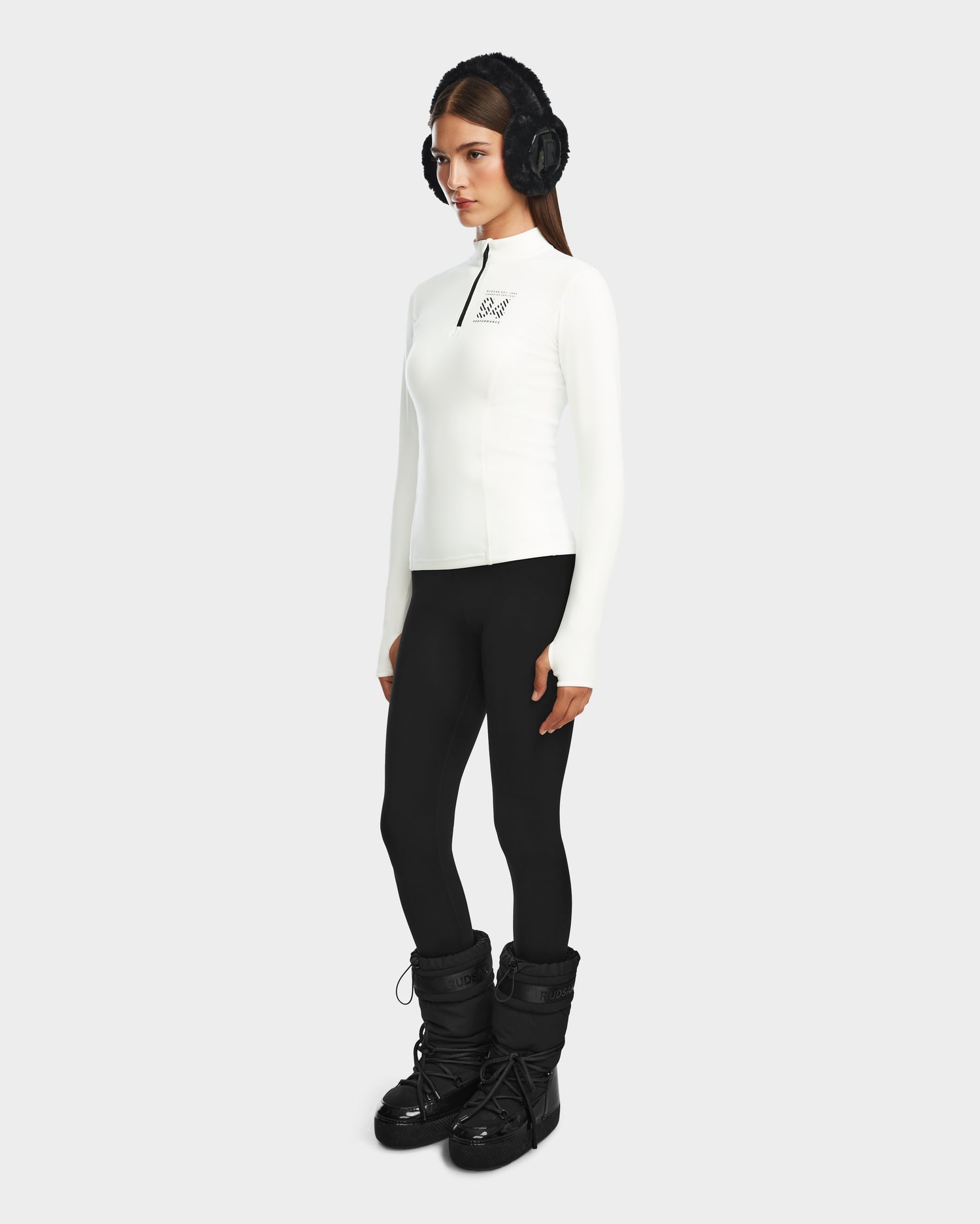 A woman stands facing forward, wearing black earmuffs and the Rudsak MARIS - 8524603 Optic White womens active top with long sleeves and a zip-up front—ideal for ski layering—against a plain light background.
