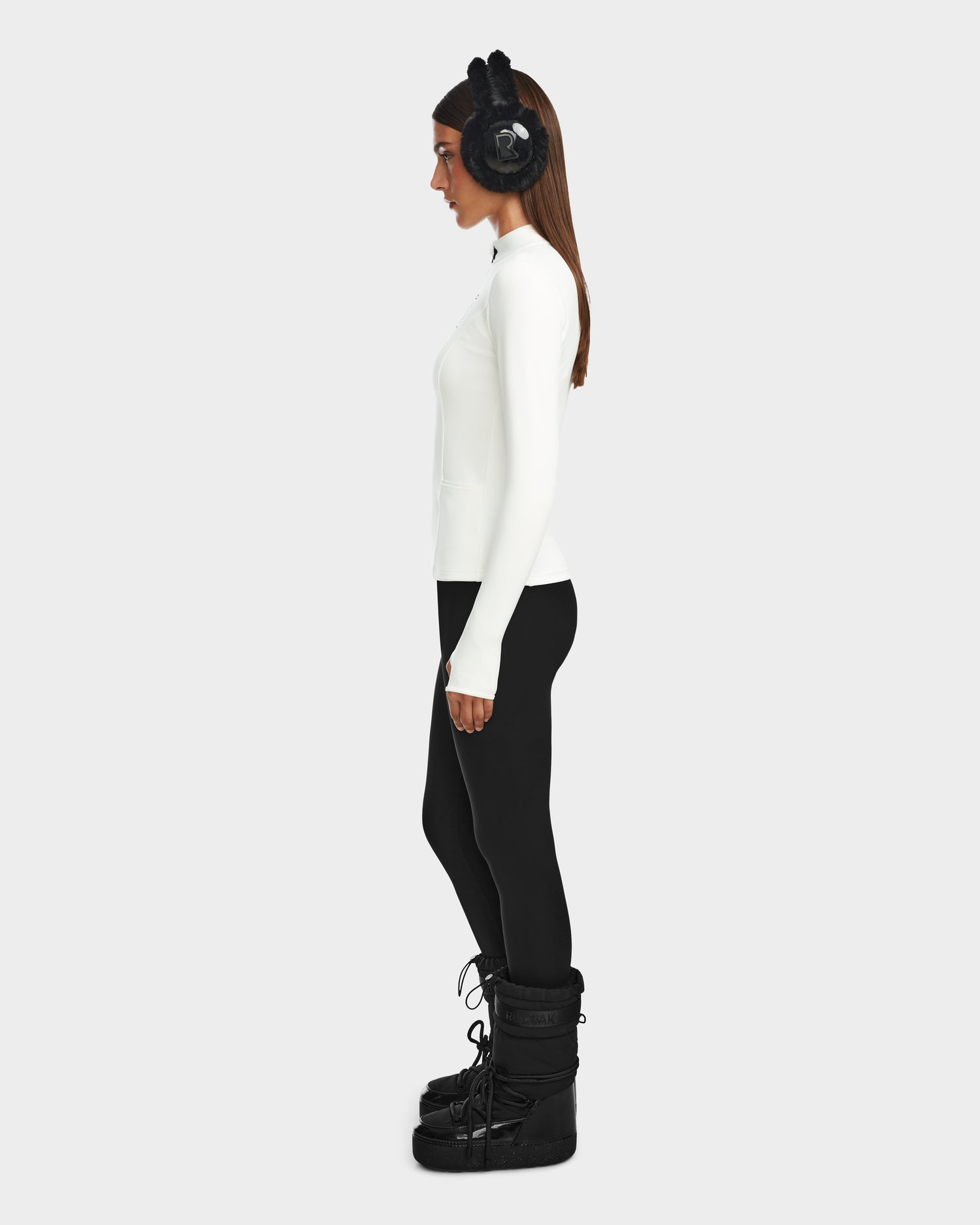 Woman in profile wears black earmuffs, black leggings, black winter boots, and the Rudsak MARIS - 8524603 Optic White women’s active top—ideal ski layering—against a plain light gray background.