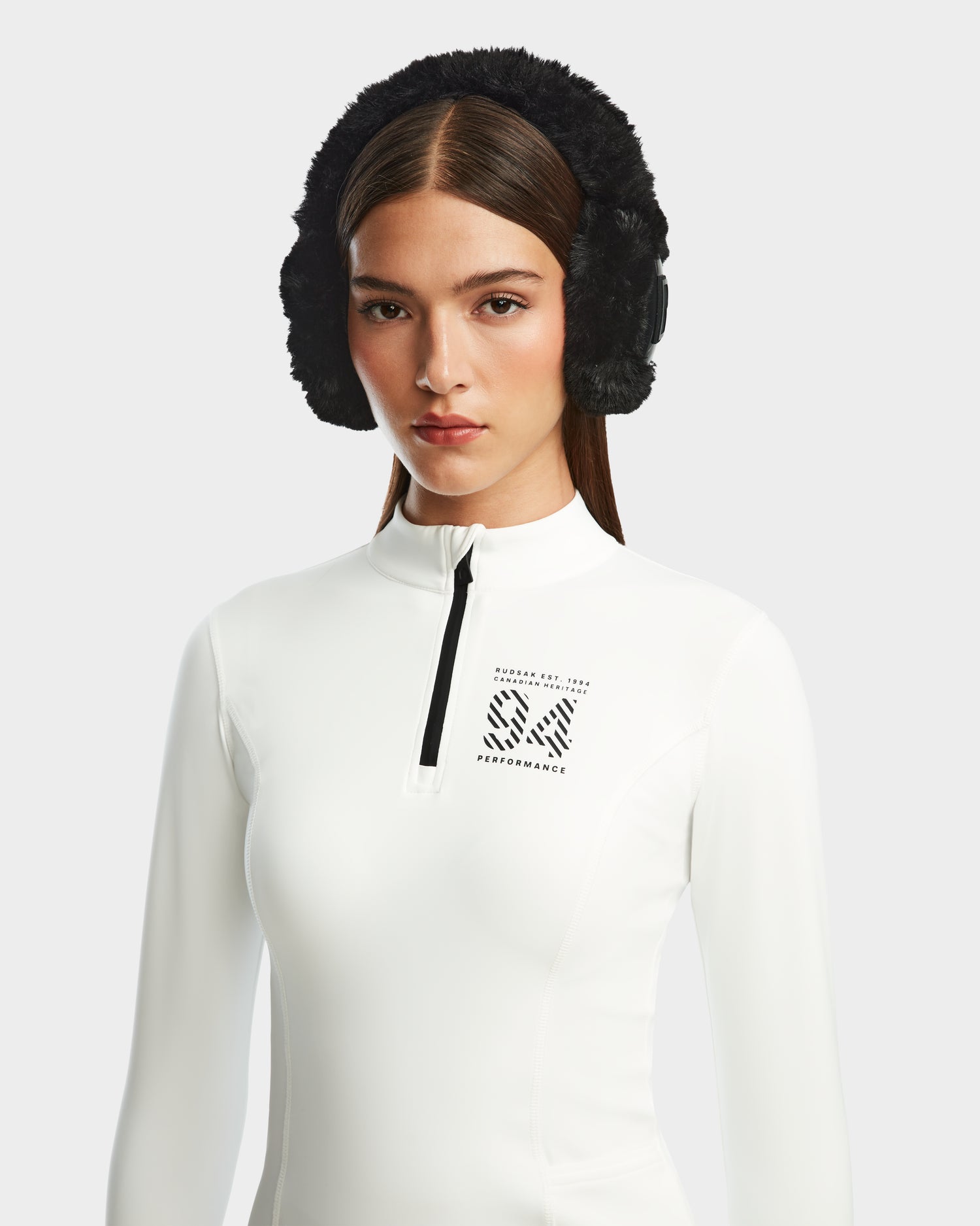 A woman with straight brown hair wears the Rudsak MARIS - 8524603 Optic White women’s active top with a black S4 PERFORMANCE logo and black earmuffs, standing against a light gray background—ideal for ski layering or winter sports.