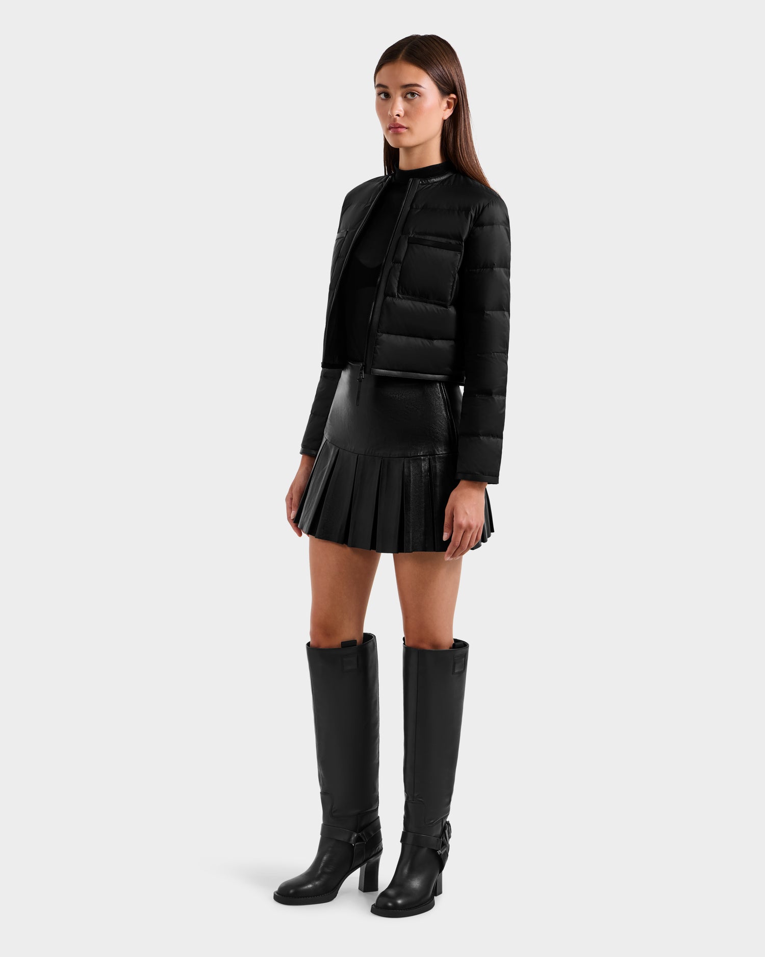A woman with long brown hair and a neutral expression wears the Rudsak MARKLE - 8125033 Black women’s cropped down jacket, paired with a black pleated mini skirt, black top, and tall heeled boots against a plain background.