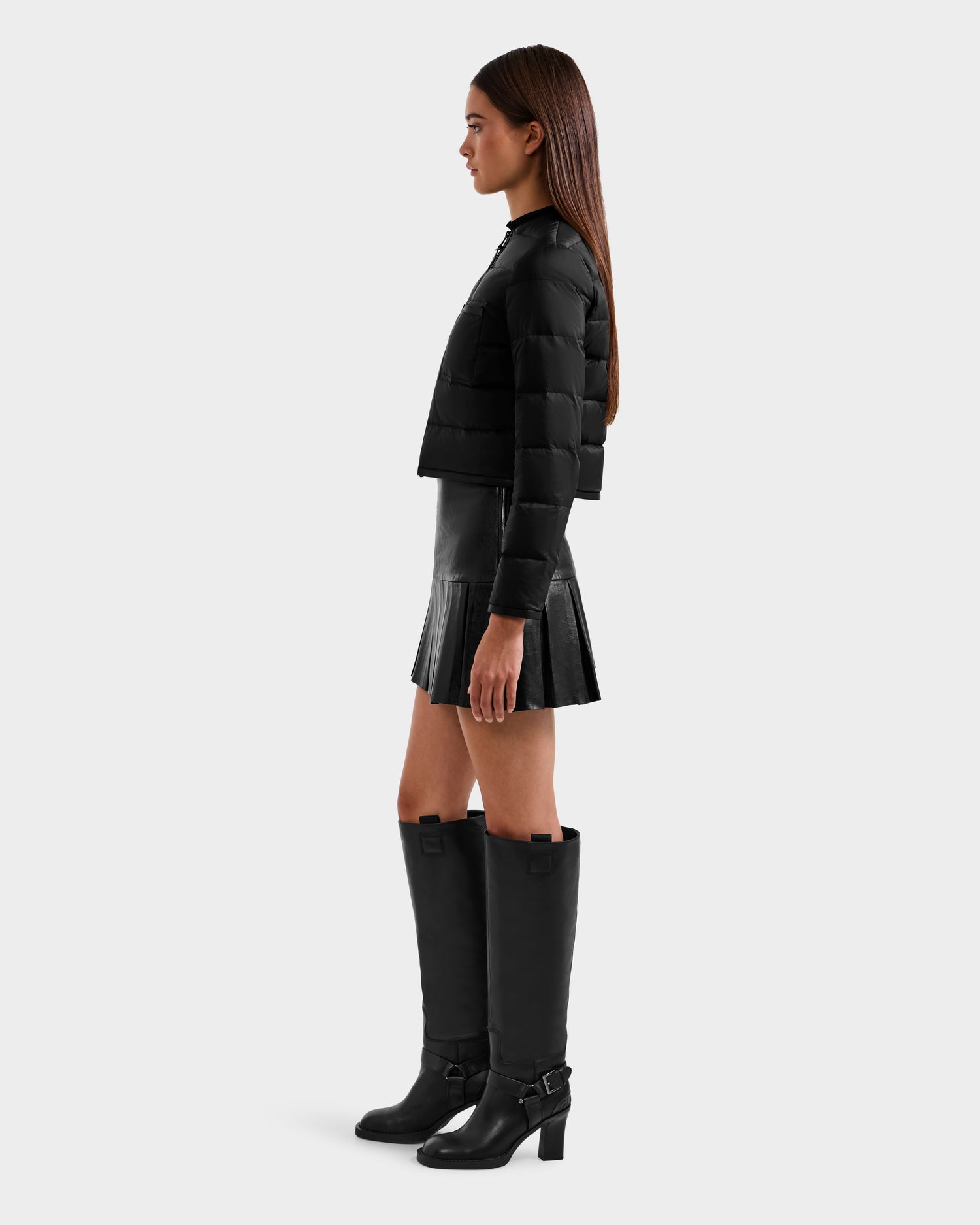 A woman in profile wears the Rudsak MARKLE - 8125033 Black cropped down jacket with a black pleated mini skirt and knee-high black buckle boots, set against a plain light gray background.