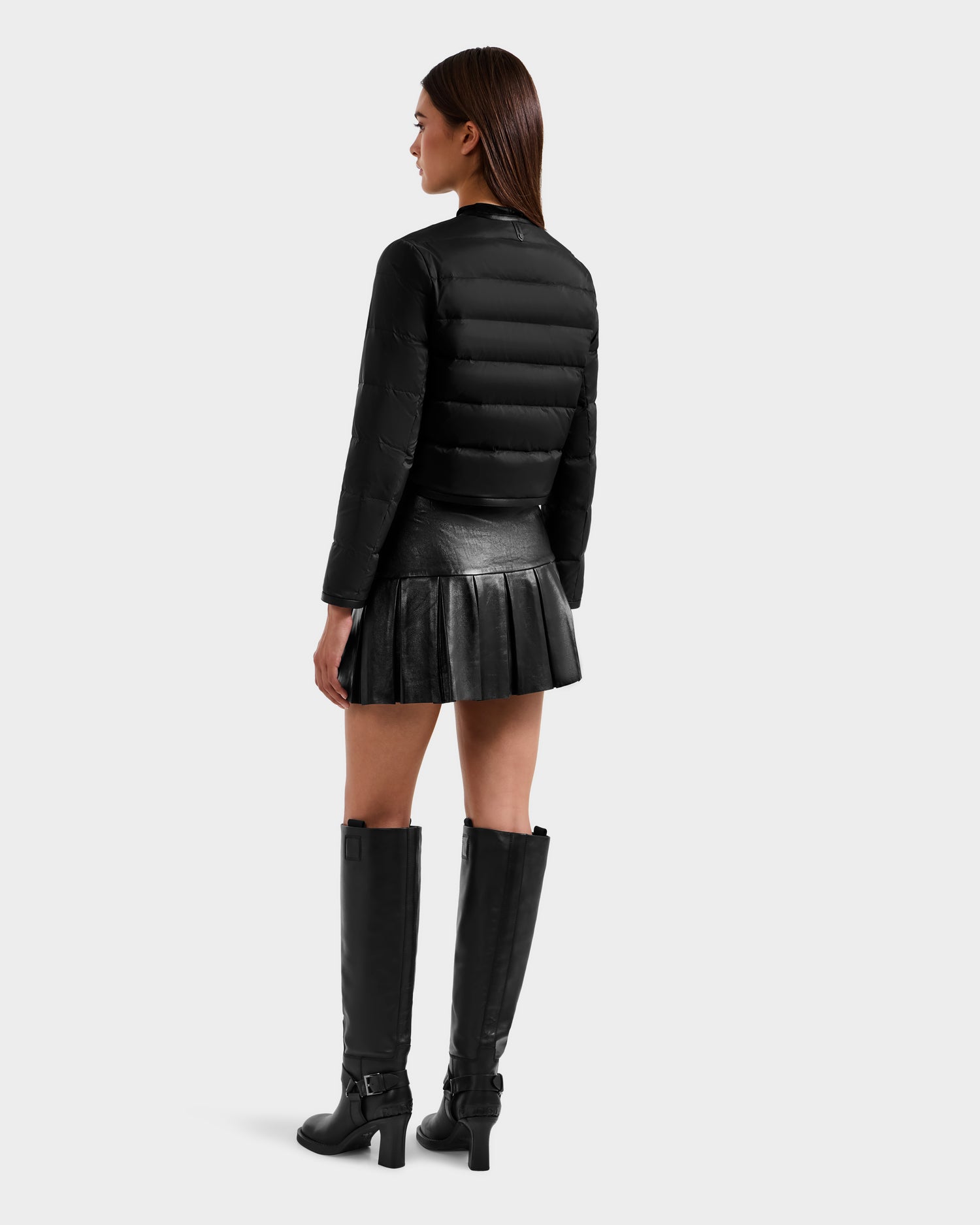 A woman with straight brown hair stands facing away, wearing the Rudsak MARKLE - 8125033 Black cropped down jacket, a black pleated leather mini skirt, and knee-high black boots with buckles against a plain light gray background.