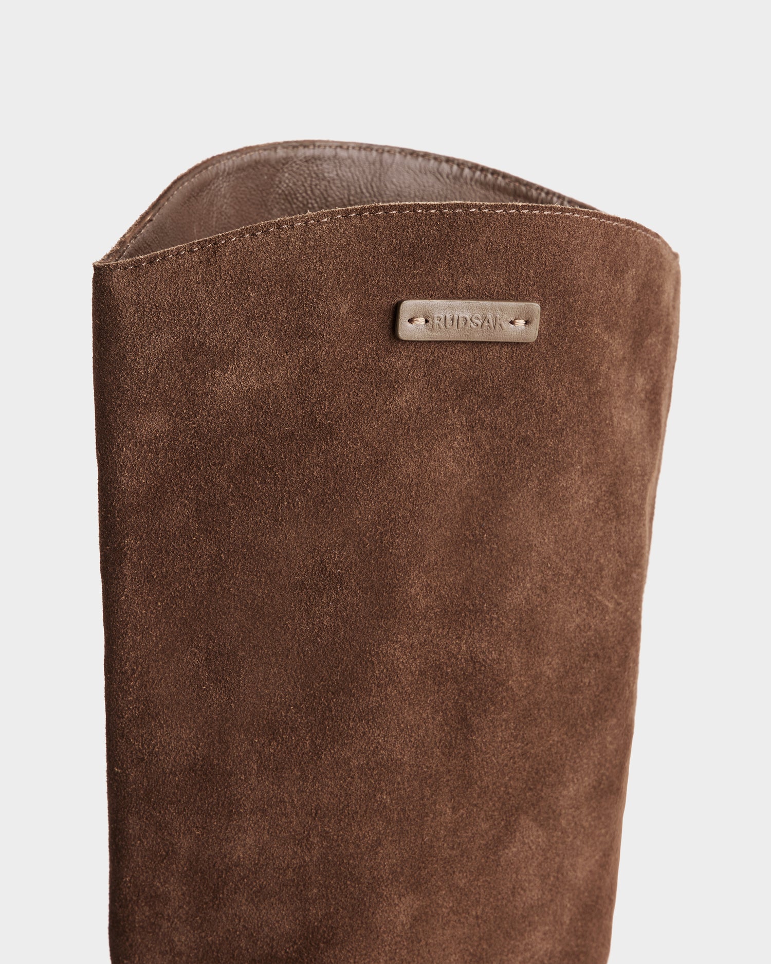 Close-up of the upper part of the Rudsak MARSALLA - 8225033 Mocha women’s suede riding boot, featuring a small metallic RUDSAK tag on the side, set against a plain white background.