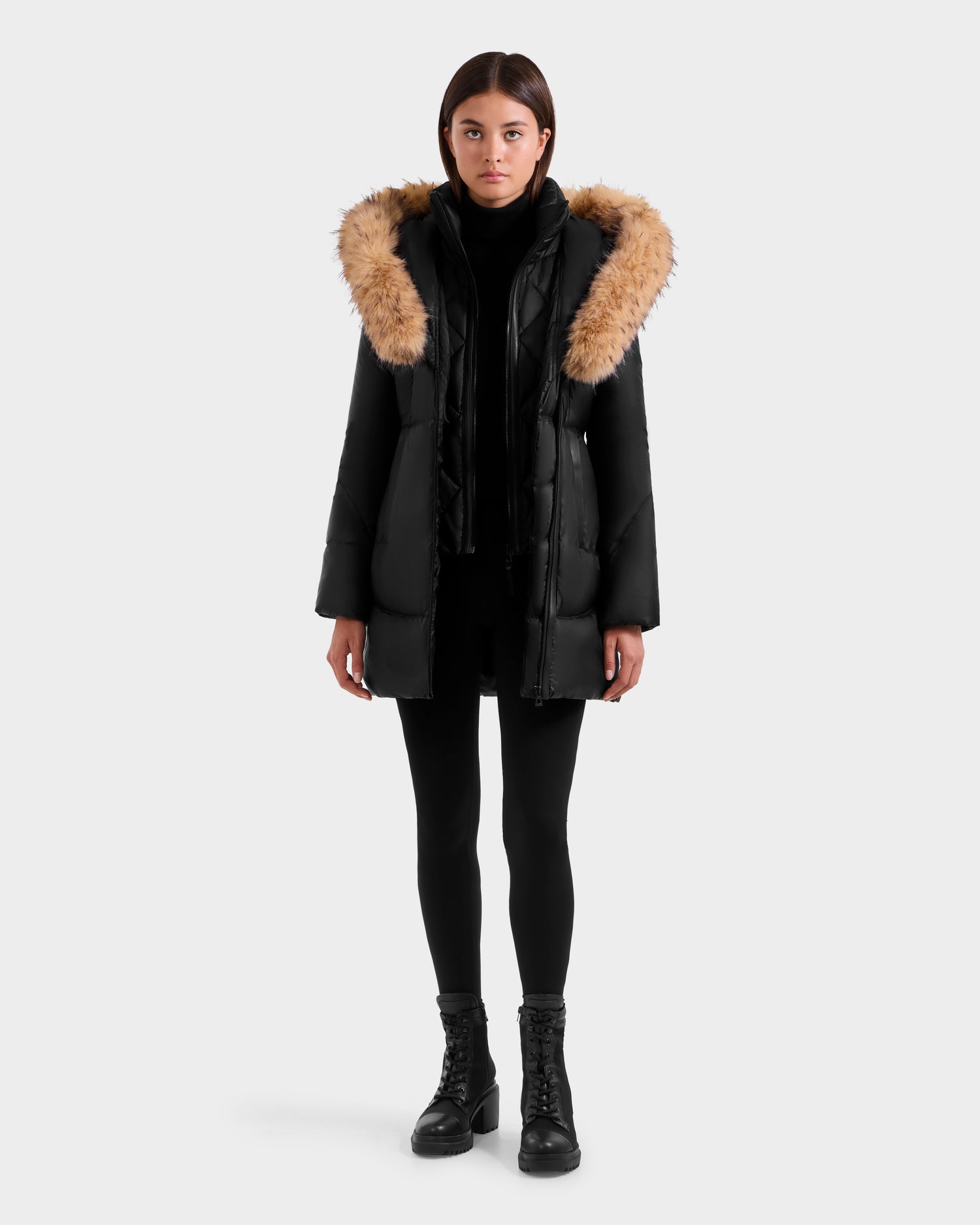 A person stands facing forward against a light gray background, wearing the Rudsak MARTINA FF - L125609 Black mid-length puffer coat with removable faux fur trim, paired with black leggings, boots, and a turtleneck.