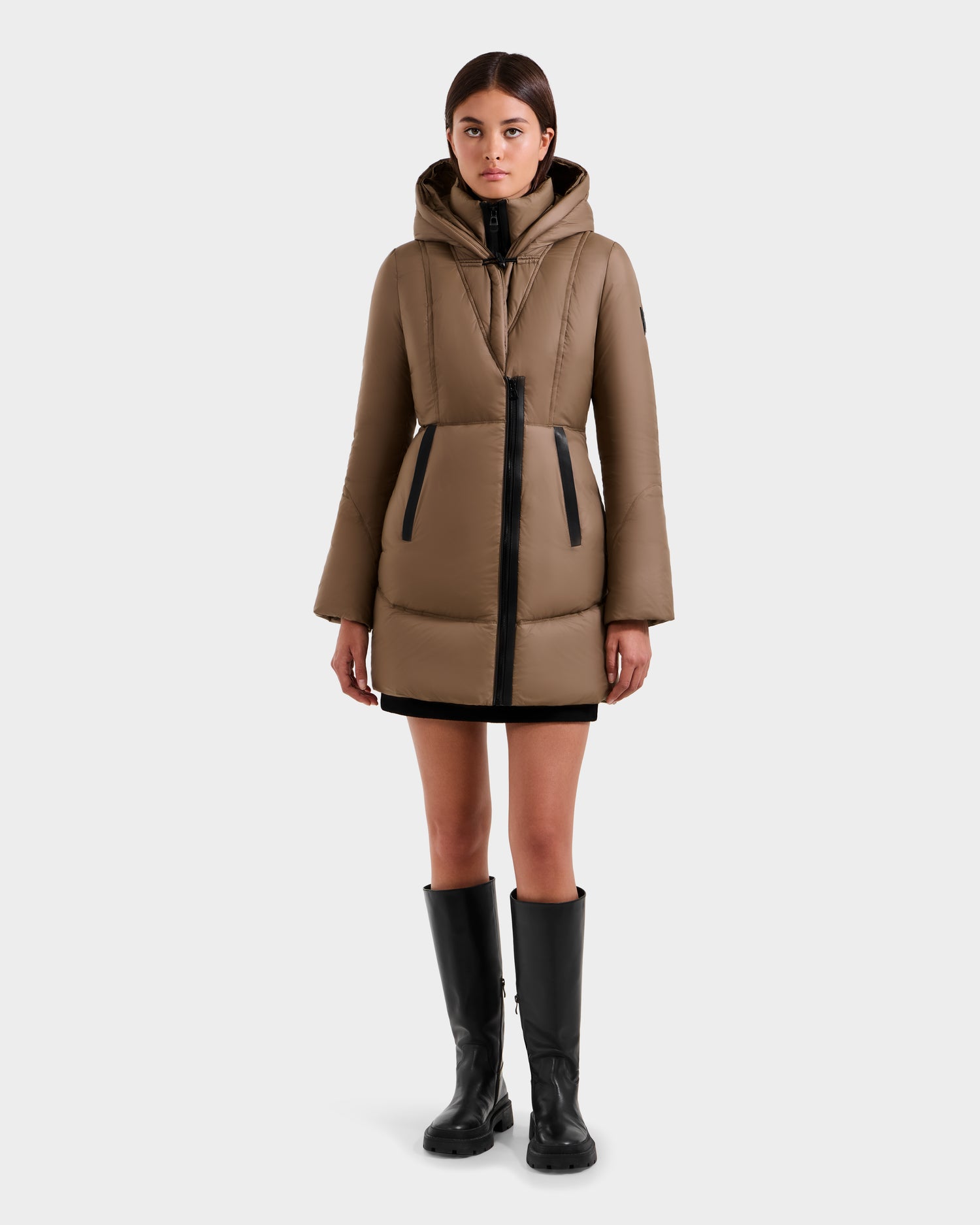 A woman faces forward in the Rudsak MARTINA FF - L125609 Taupe mid-length puffer coat with removable faux fur trim, black zippers, a black mini skirt, and knee-high black boots, set against a plain white background.