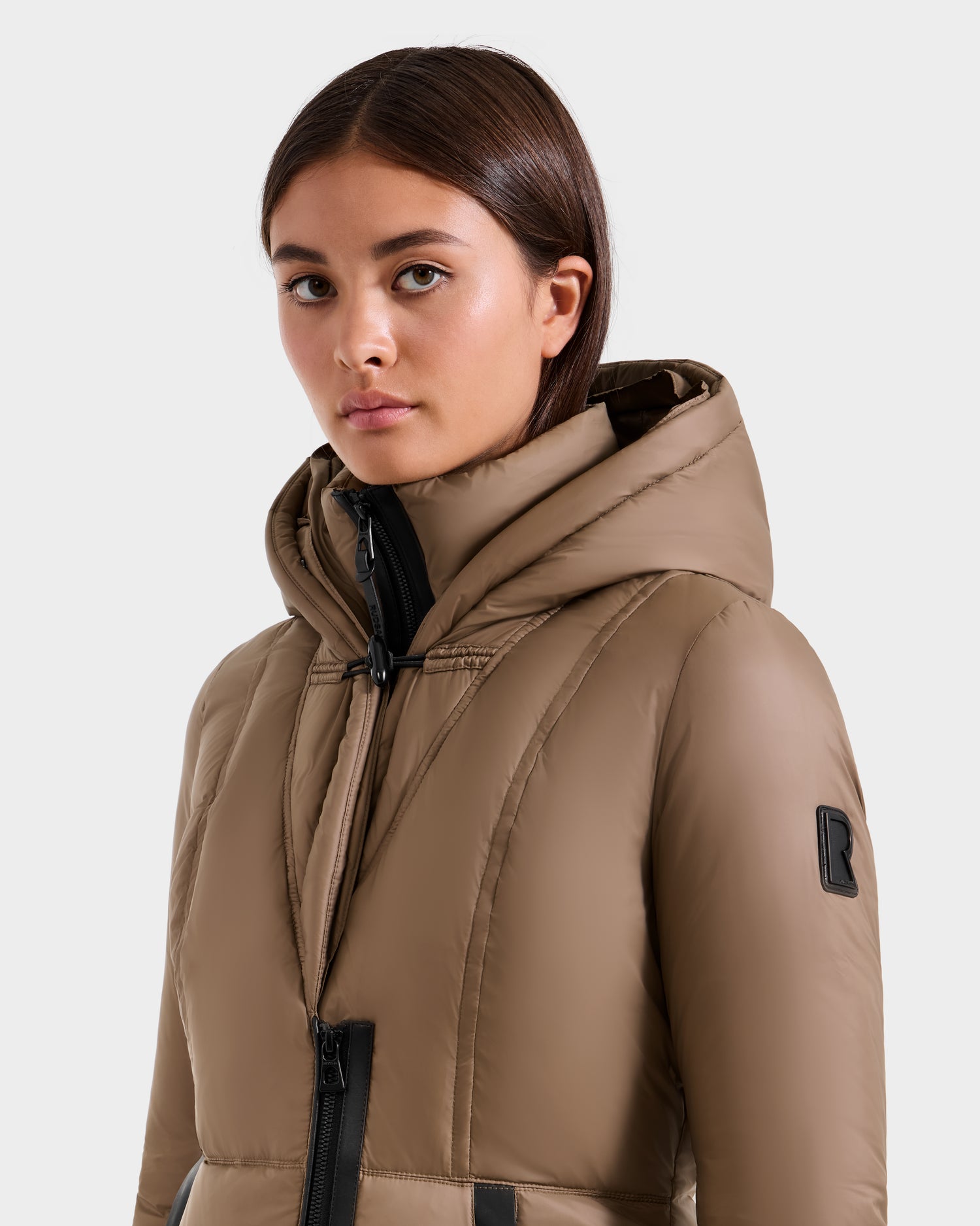 A woman with straight brown hair wears the Rudsak MARTINA FF - L125609 Taupe mid-length puffer jacket with black zippers, looking slightly to the side against a plain light background.