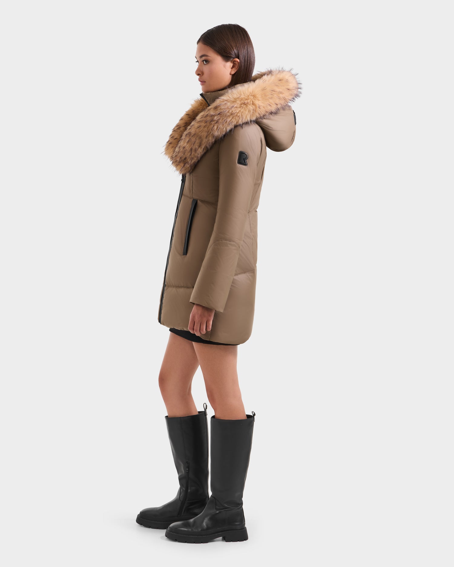 A woman in profile wears the Rudsak MARTINA FF - L125609 Taupe, a mid-length taupe puffer with a removable faux fur-trimmed hood and black zipper pockets, paired with knee-high black boots against a plain white background.