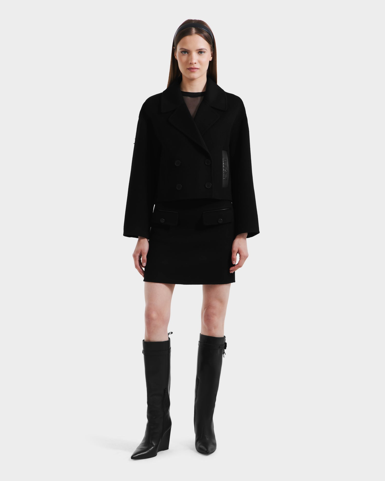 A woman stands facing forward wearing the Rudsak MARTINEZ - 8125580 Black vegan leather cropped double-breasted jacket, paired with a black mini skirt and knee-high black boots against a plain light background.