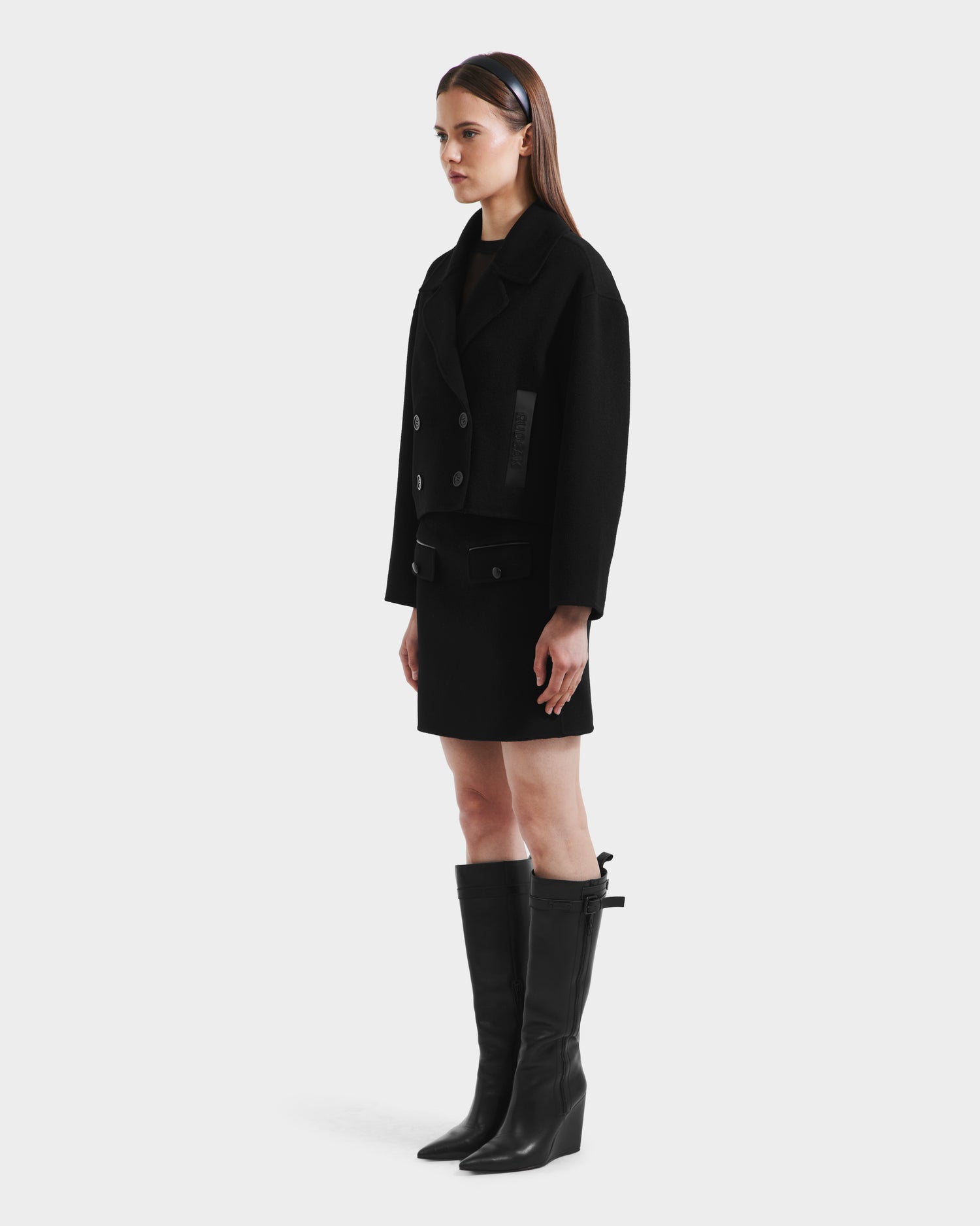 A woman stands in profile against a white background, wearing the Rudsak MARTINEZ - 8125580 Black double-breasted wool-blend jacket, paired with a black skirt, knee-high boots, and a headband.