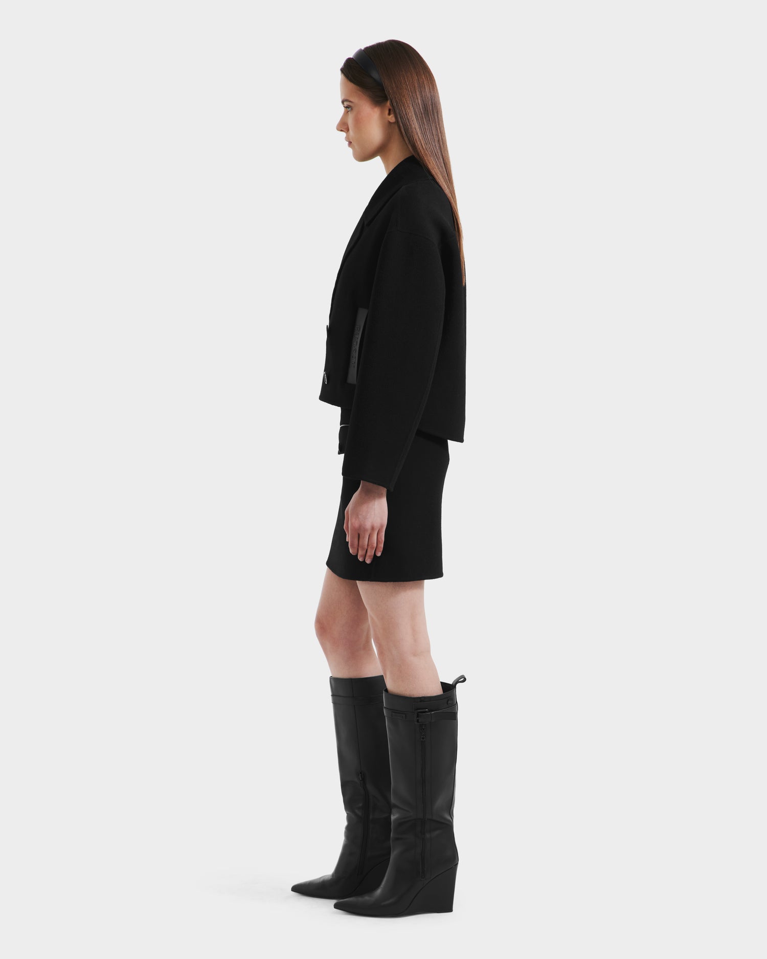 A woman stands in profile against a plain background, wearing the Rudsak MARTINEZ - 8125580 Black blazer with a matching skirt and knee-high black boots. Her straight hair is down, and her arms rest relaxed by her sides.