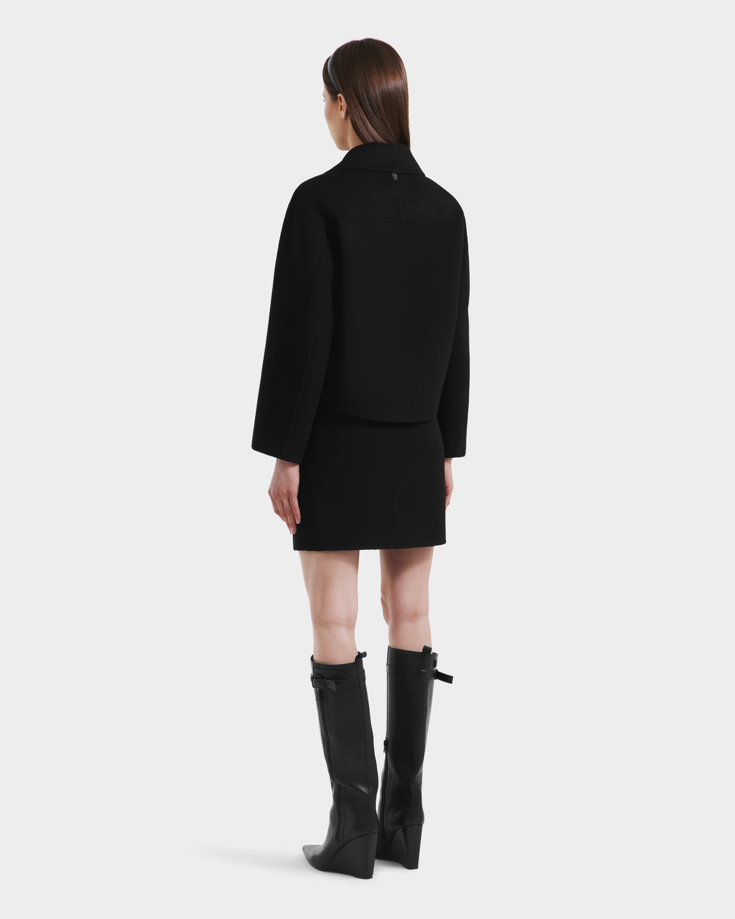 A woman with straight brown hair stands facing away, wearing the Rudsak MARTINEZ - 8125580 Black vegan leather jacket, a black skirt, and knee-high black boots against a plain white background. Her arms rest relaxed at her sides.