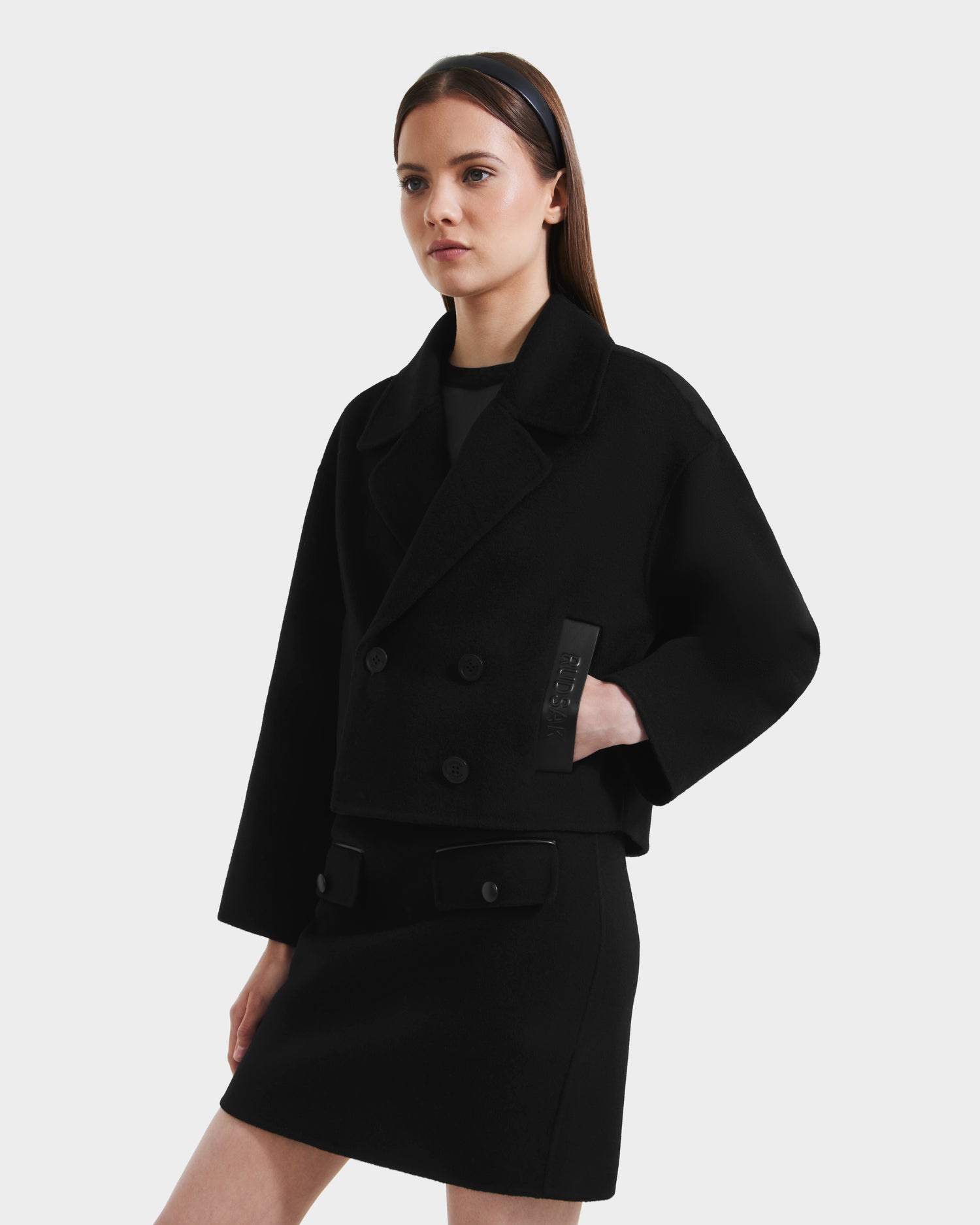 A woman wearing the Rudsak MARTINEZ - 8125580 Black vegan leather double-breasted jacket with wide lapels and a matching black skirt stands against a white background, looking to the side with one hand in her pocket.