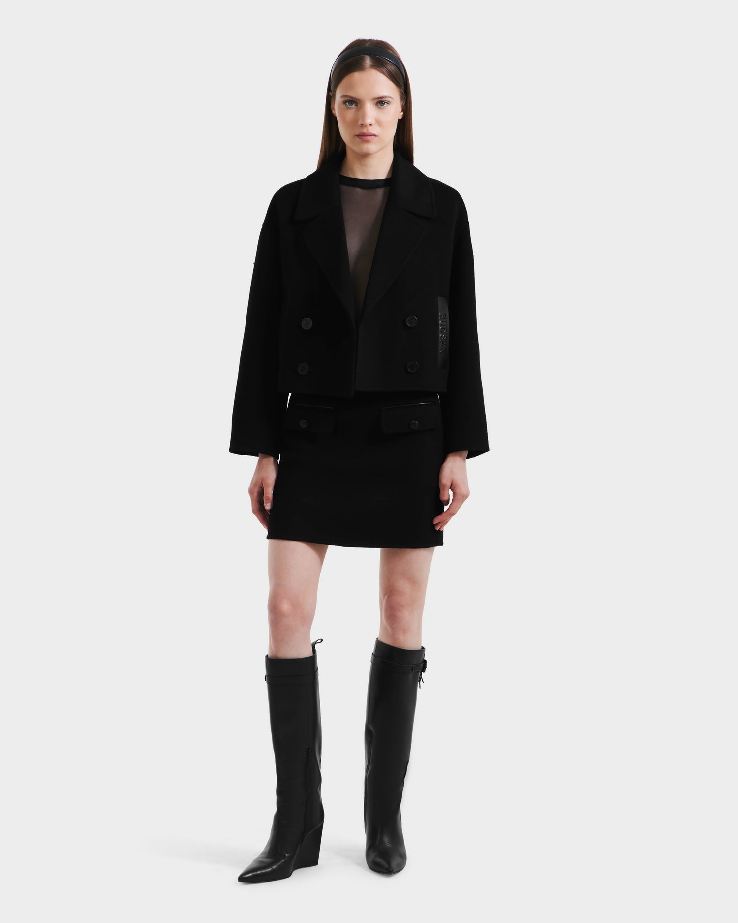 A woman with straight brown hair and a neutral expression wears the Rudsak MARTINEZ - 8125580 Black vegan leather mini skirt, a black cropped jacket, knee-high boots, and a sheer black top, standing against a plain white background.