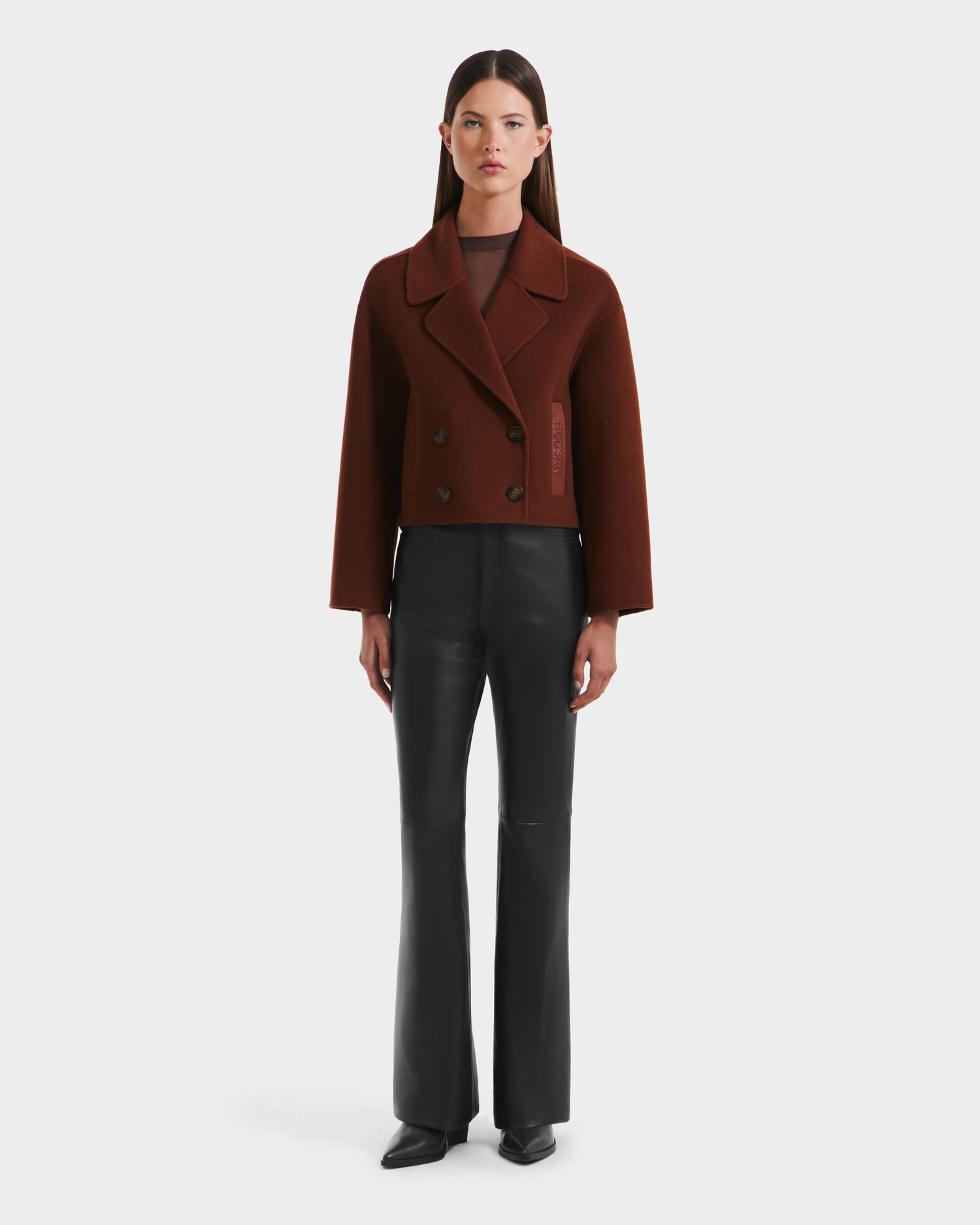 A woman with straight brown hair and a neutral expression stands against a plain background wearing the Rudsak MARTINEZ - 8125580 Rust women’s short coat, black vegan leather pants, and black shoes.