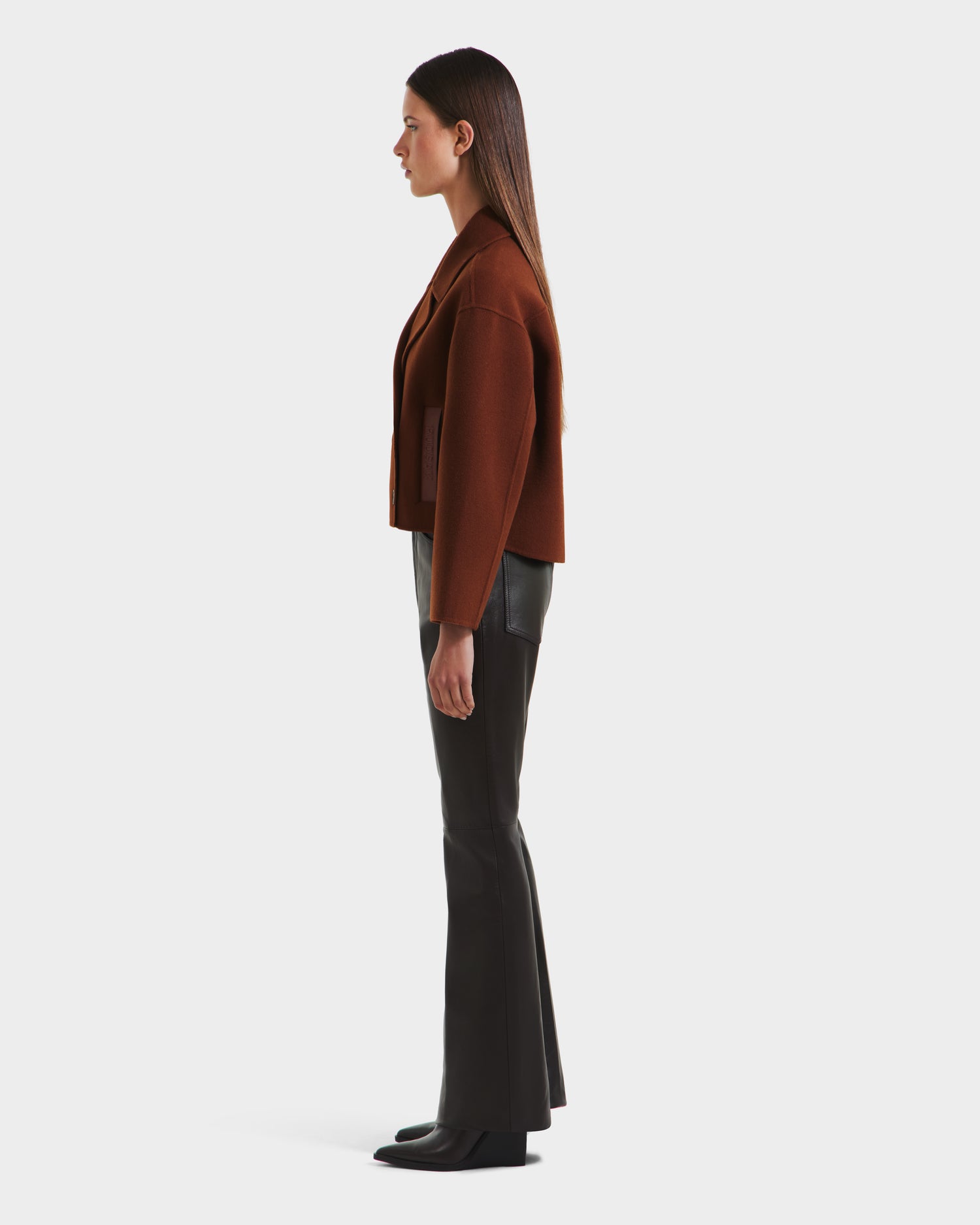A woman in profile faces left against a white background, wearing the Rudsak MARTINEZ - 8125580 Rust wool-blend oversized jacket, black flared pants, and black shoes. Her straight hair is worn down.