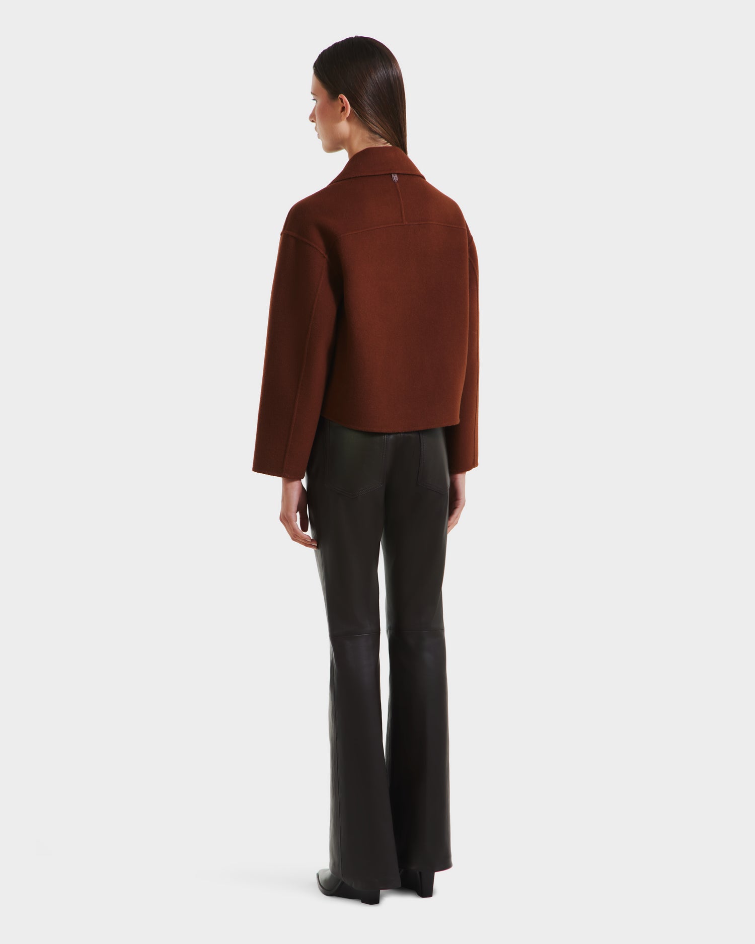 A woman faces away, wearing the MARTINEZ - 8125580 Rust jacket by Rudsak and black flared pants against a plain white background.