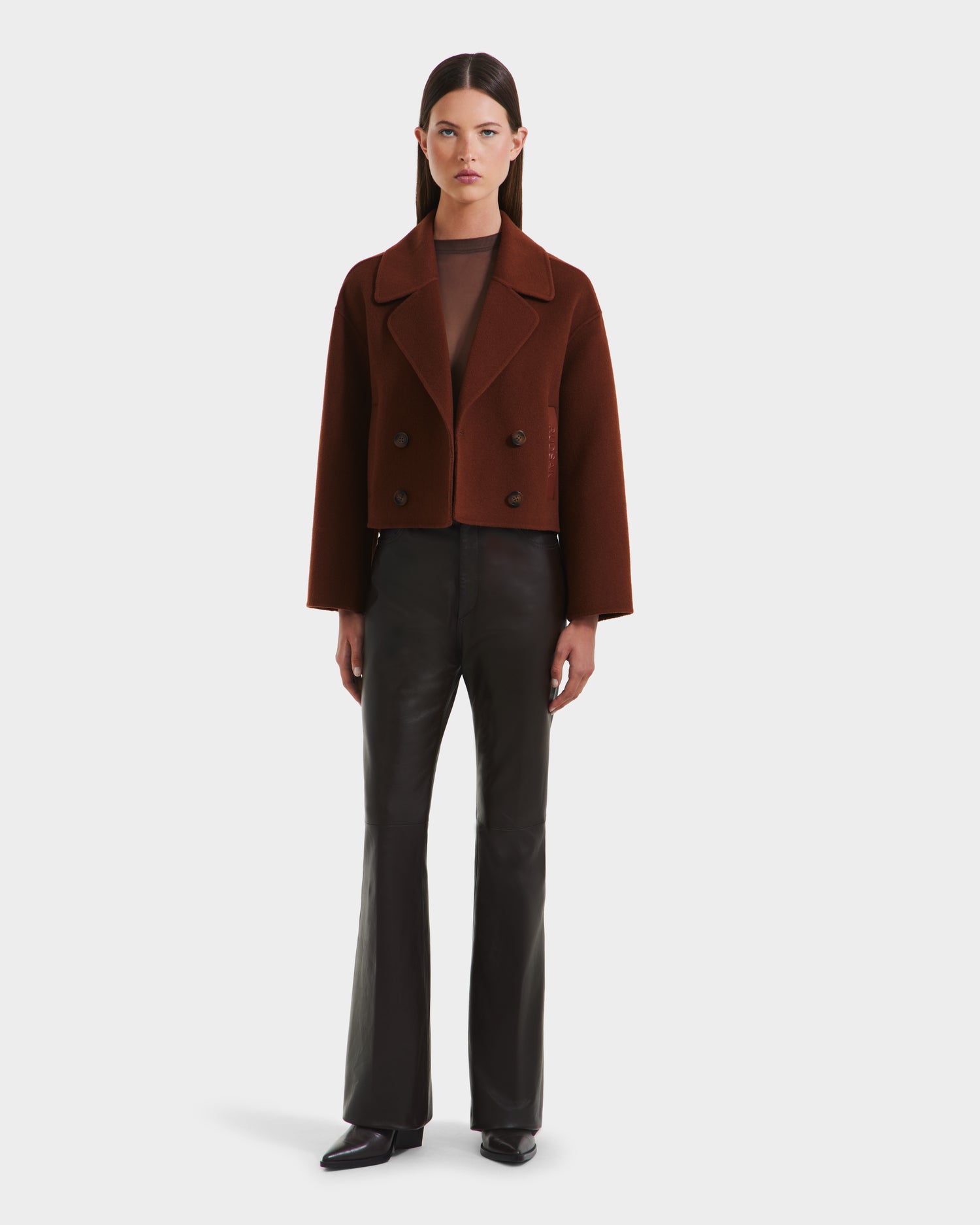 A woman faces forward wearing the Rudsak MARTINEZ - 8125580 Rust cropped double-breasted short coat over a sheer brown top and high-waisted black vegan leather pants, with black pointed-toe shoes, against a plain white background.