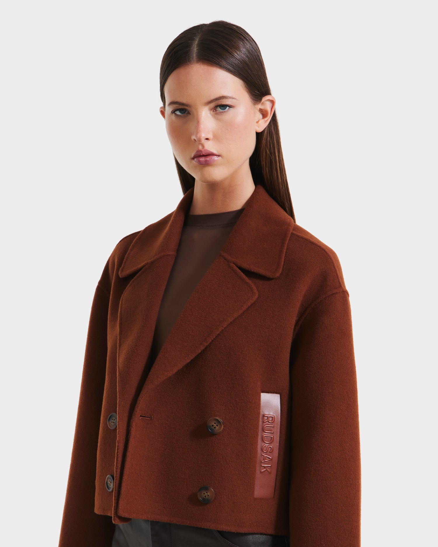 A woman with straight brown hair wears the Rudsak MARTINEZ - 8125580 Rust womens short wool-blend double-breasted coat with wide lapels and a signature patch, layered over a sheer brown top, standing against a plain light background.