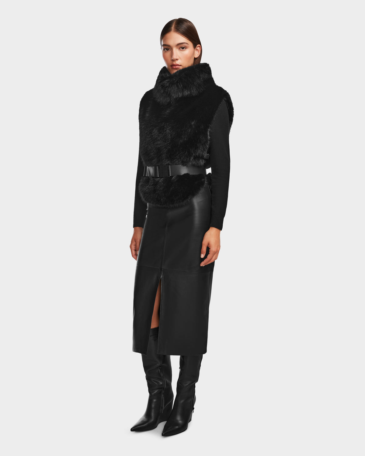 A woman wears the Rudsak MAULIN - 8524611 Black vegan leather midi skirt with a front slit, styled with a black faux fur vest, mock-neck pullover, and knee-high boots against a plain, light background.
