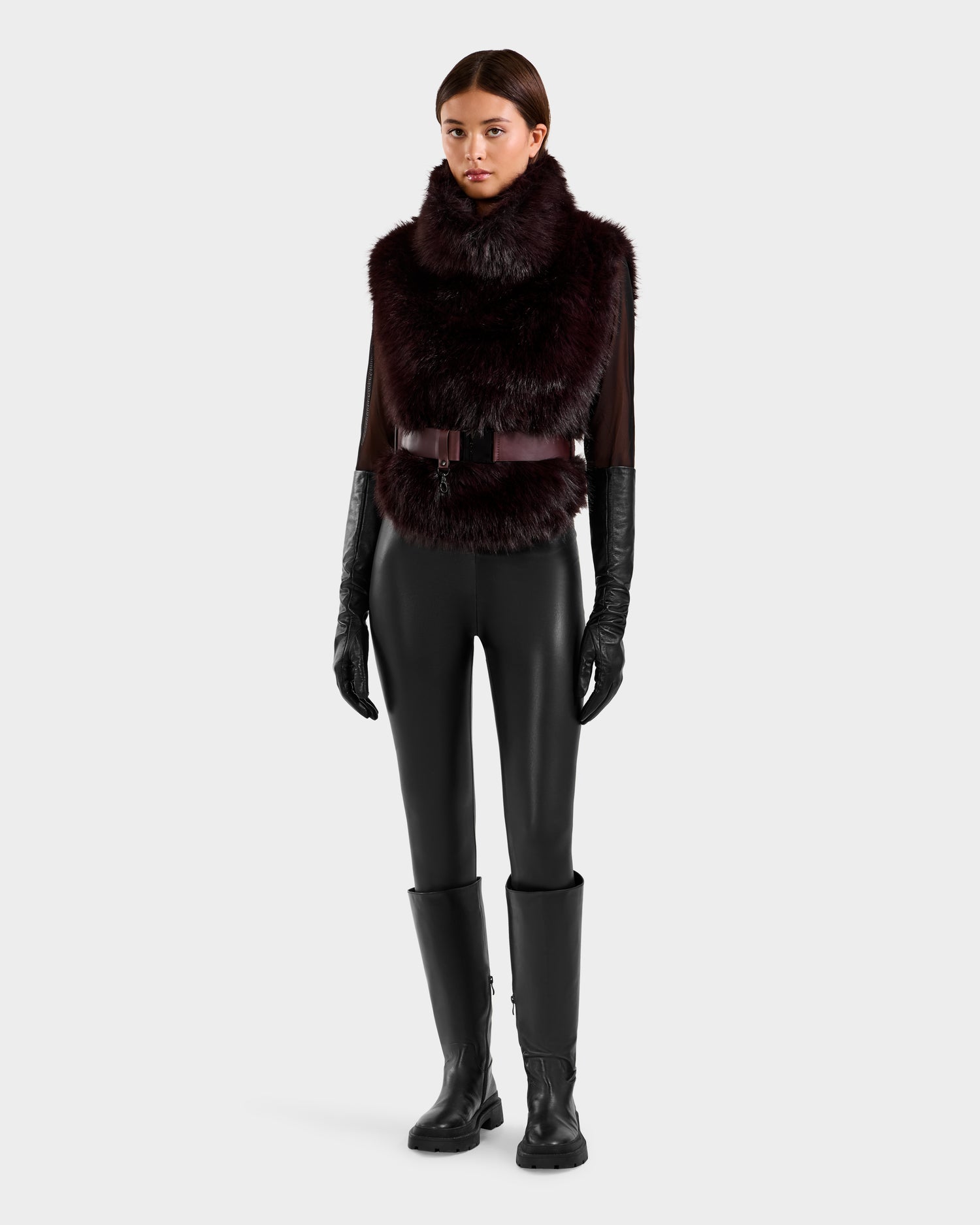 A woman faces forward in the Rudsak MAULIN - 8524611 Bordeaux, a faux fur vest with a high collar, styled with black leather gloves, fitted pants, a wide belt, and knee-high black boots against a plain light background.