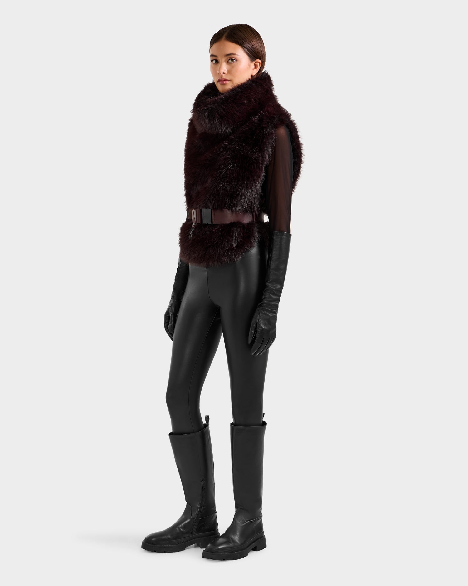 A woman models the Rudsak MAULIN - 8524611 Bordeaux, a large dark faux fur vest, paired with a black belted top, leather gloves, fitted pants, and knee-high boots against a plain white background.