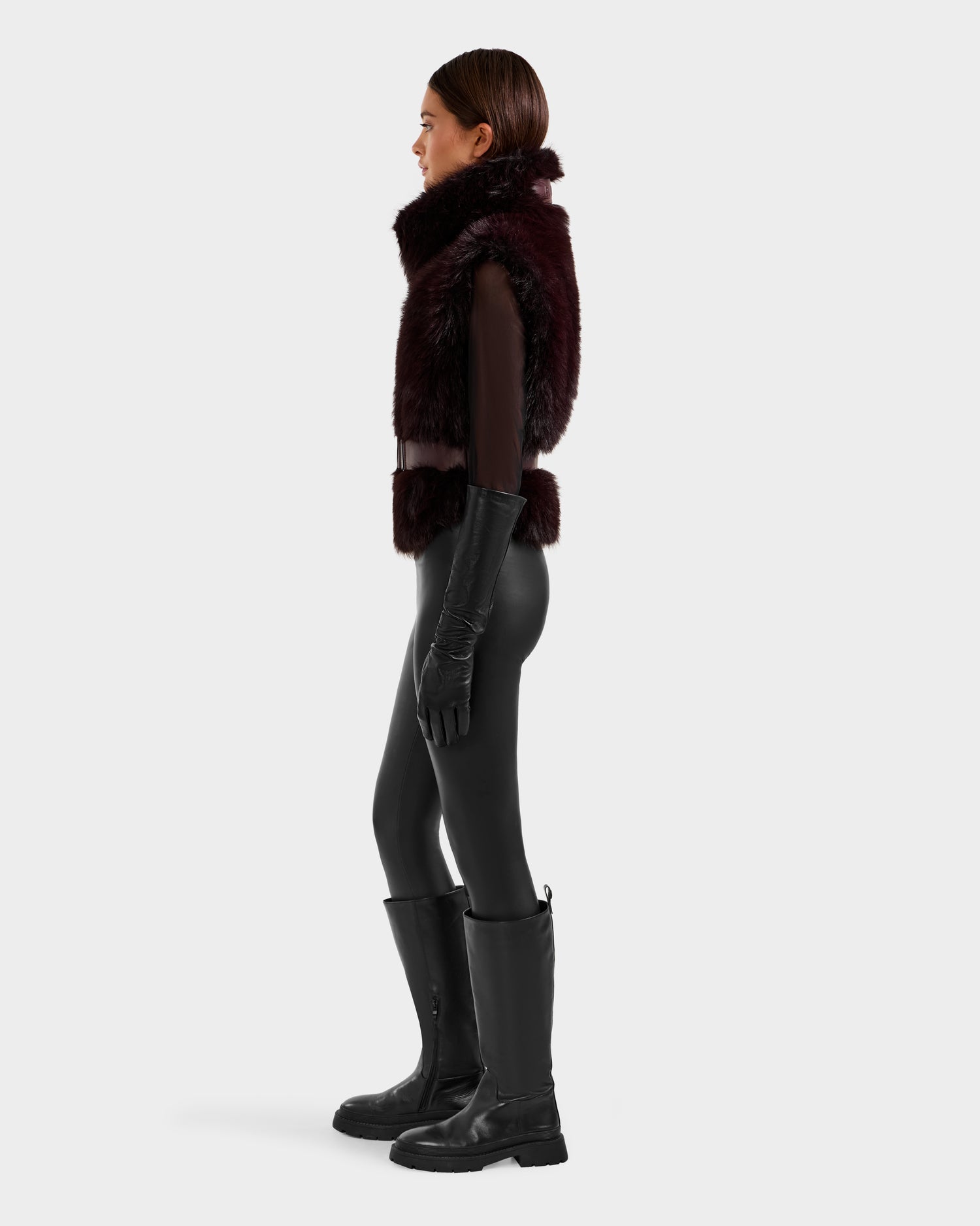 A person stands in profile against a plain light background, wearing the Rudsak MAULIN - 8524611 Bordeaux faux fur vest with a sheer long-sleeve top, black gloves, shiny leggings, and knee-high black boots.