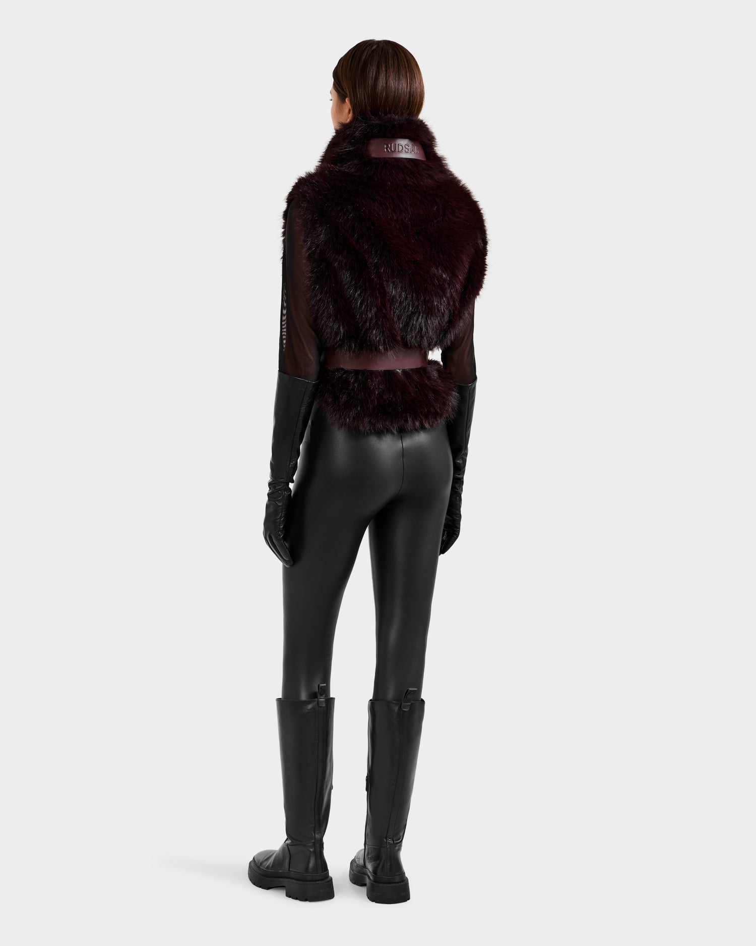 A person faces away, wearing the Rudsak MAULIN - 8524611 Bordeaux vegan leather vest over a sheer black top, with black gloves, shiny pants, and knee-high boots against a plain light background.