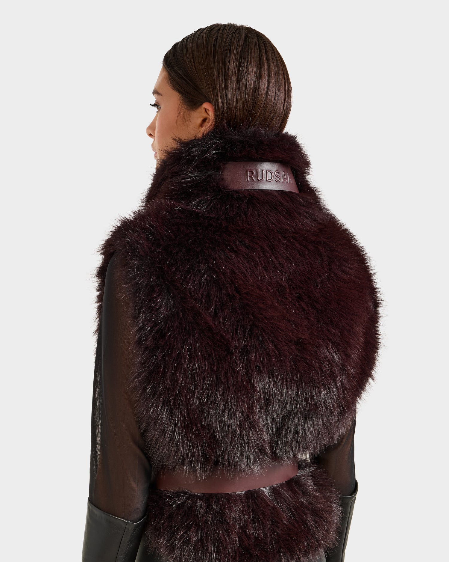 A woman with straight brown hair models the Rudsak MAULIN - 8524611 Bordeaux faux fur vegan leather vest, featuring “RUDSAK” on the high collar. She wears it over a sheer black long-sleeve shirt, showing the vest’s back.