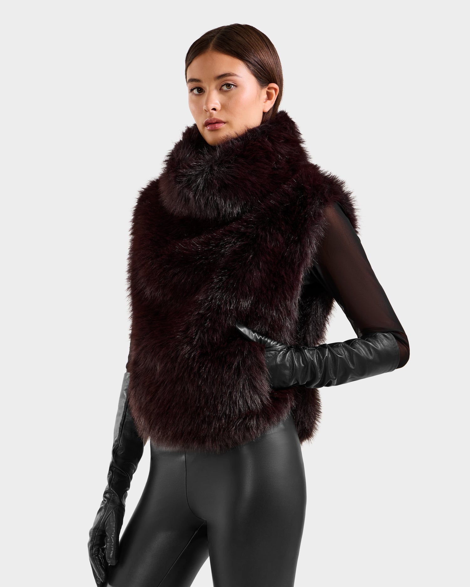 A woman models the Rudsak MAULIN - 8524611 Bordeaux sleeveless faux fur vest with a high collar, paired with long black leather gloves and fitted pants, standing against a light background and highlighting its chic appeal.