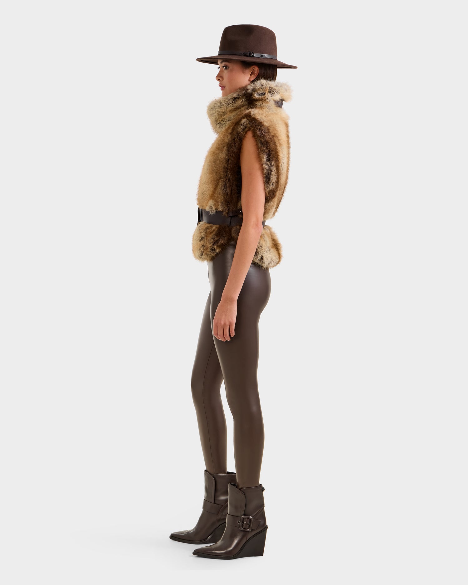 A woman models the Rudsak MAULIN - 8524611 Mix Mocha in profile, wearing a brown wide-brim hat, sleeveless faux fur vest, dark leggings, and brown ankle boots against a plain white background.