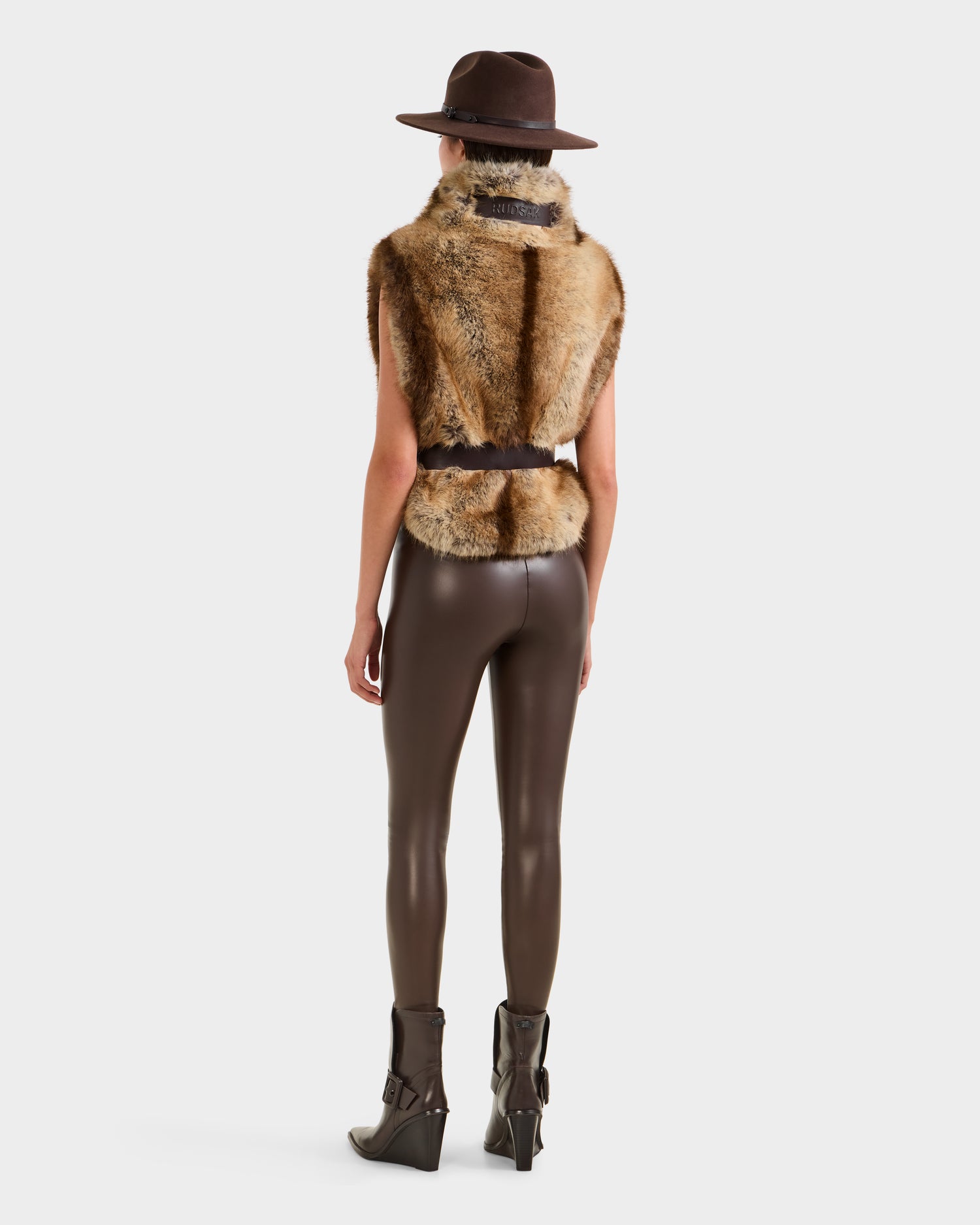 A person stands facing away, wearing Rudsak's MAULIN - 8524611 Mix Mocha: a brown wide-brim hat, sleeveless faux fur vest over brown shiny vegan leather leggings, and high-heeled ankle boots against a plain white background.