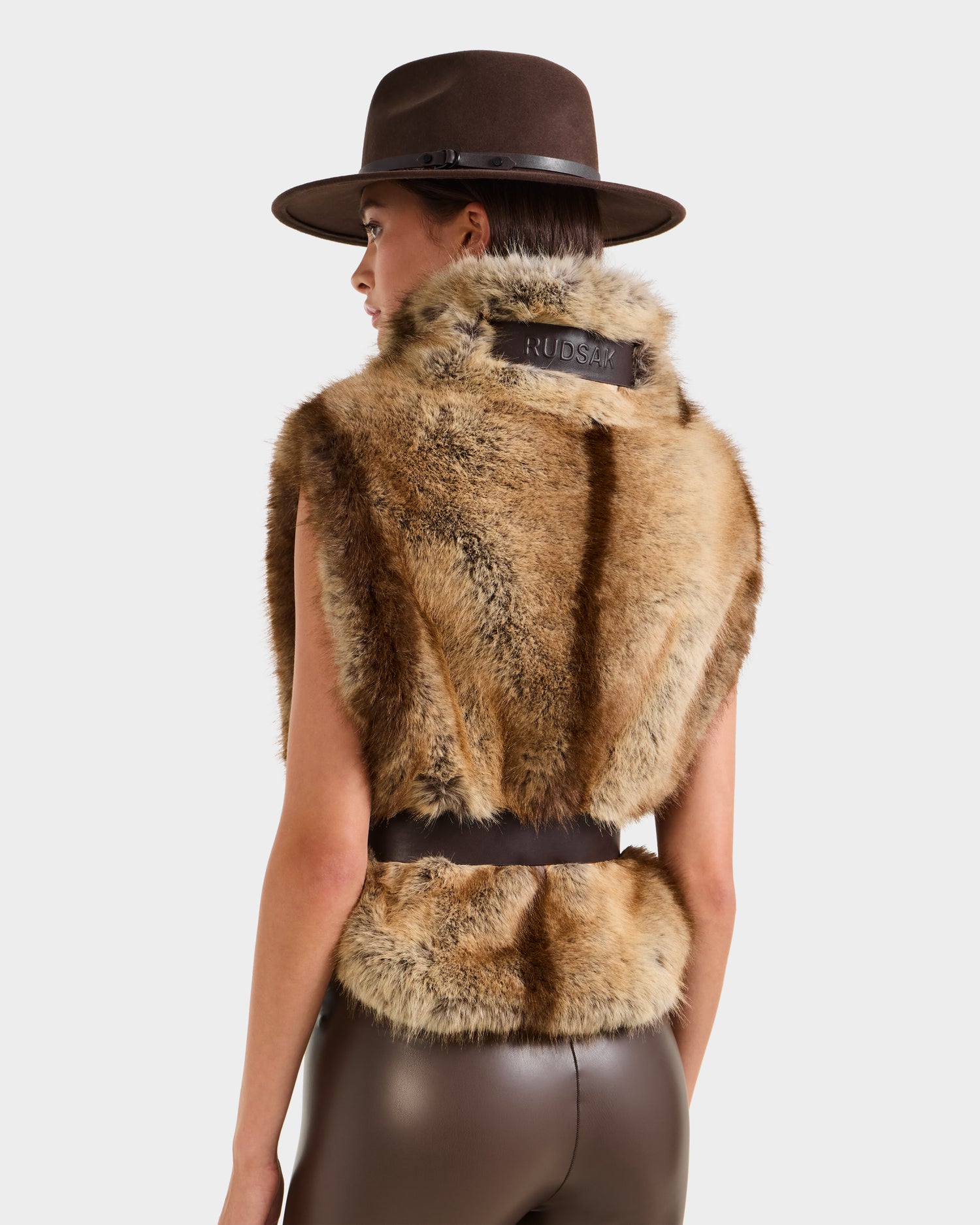 A woman is seen from behind wearing Rudsak’s MAULIN - 8524611 Mix Mocha: a brown wide-brimmed hat, sleeveless faux fur vest with vegan leather patch, and brown vegan leather pants, set against a plain background.